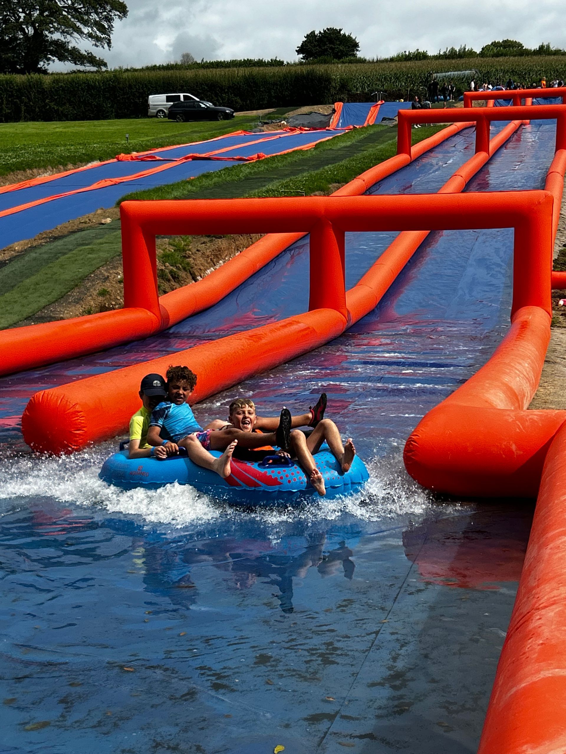 Two people slide down a long, blue inflatable water slide with orange side barriers on a sunny day.
