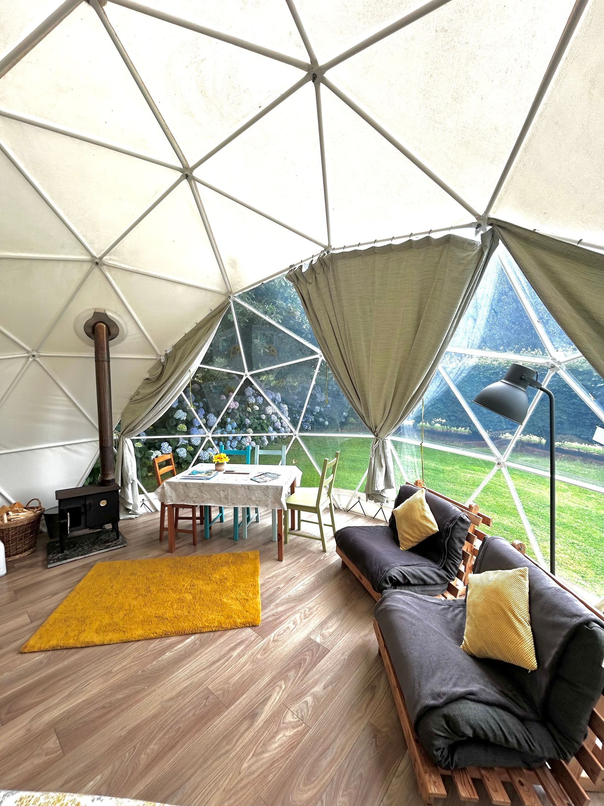 A geodesic dome glamping tent featuring a wood stove, dining table, and dark gray sofas with yellow cushions.
