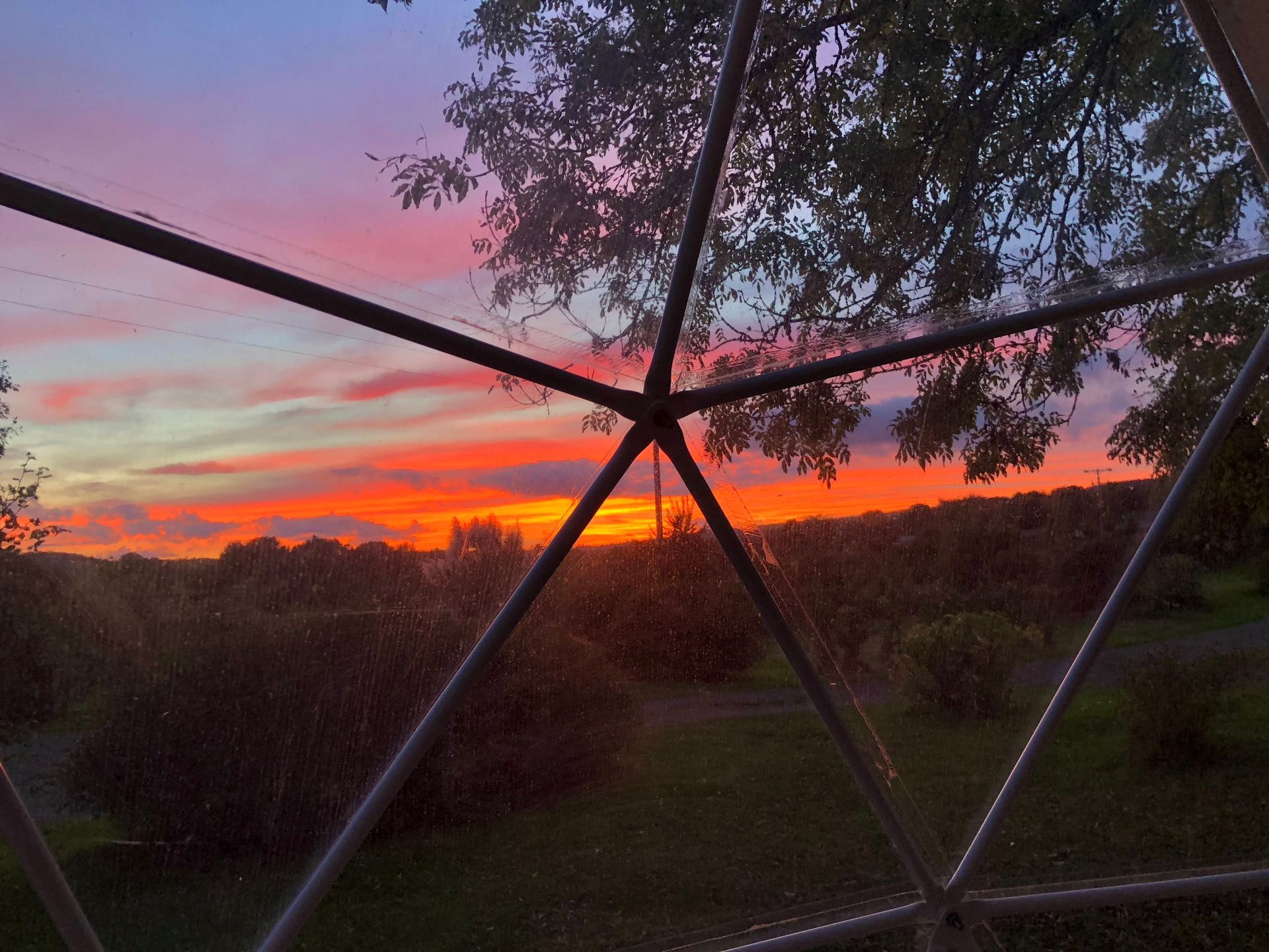 Vibrant orange and purple sunset viewed from inside a geodesic dome structure with metal struts framing the sky.