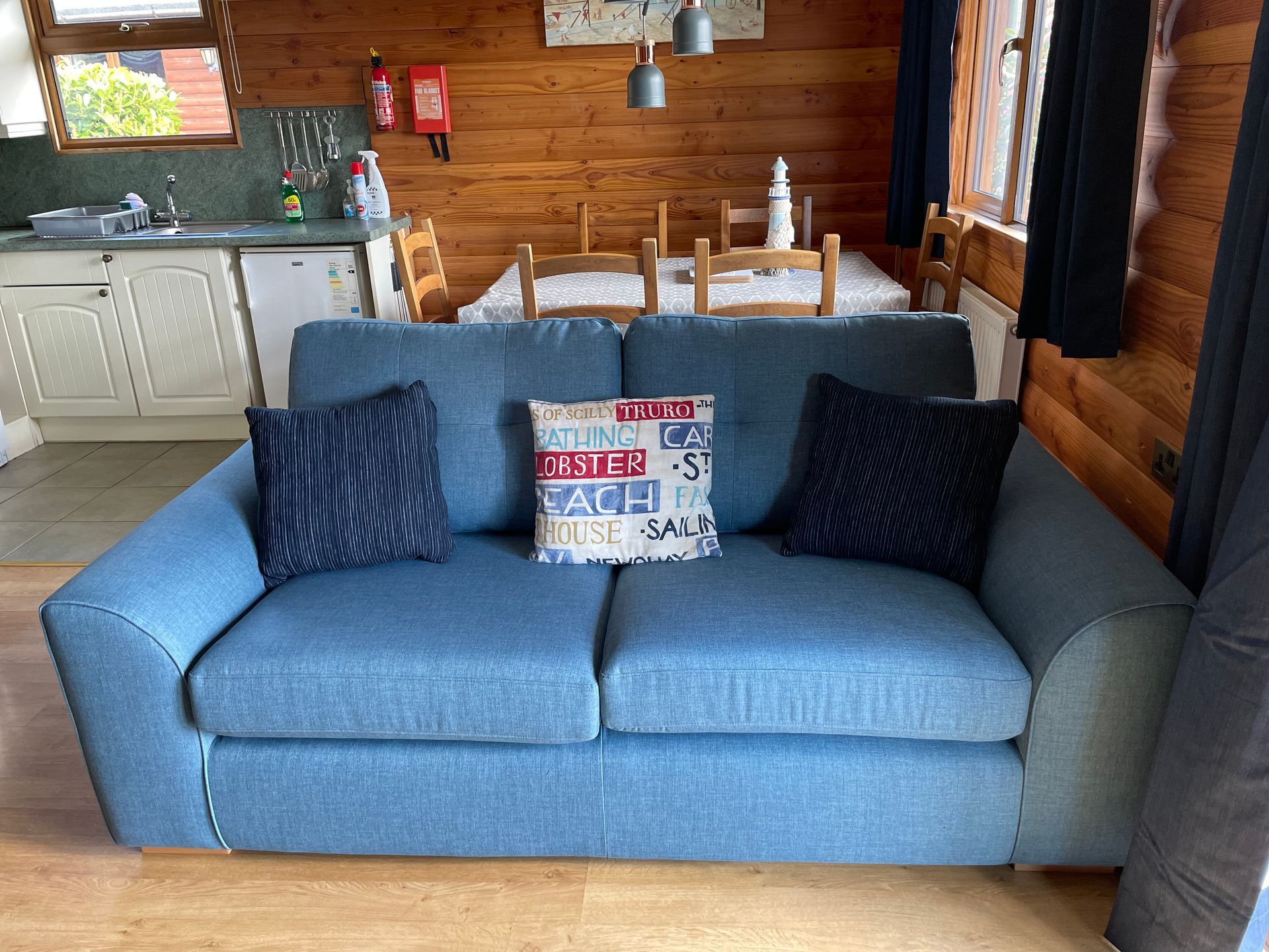 A blue two-seater sofa with three cushions placed in front of a dining table in a rustic wooden room.