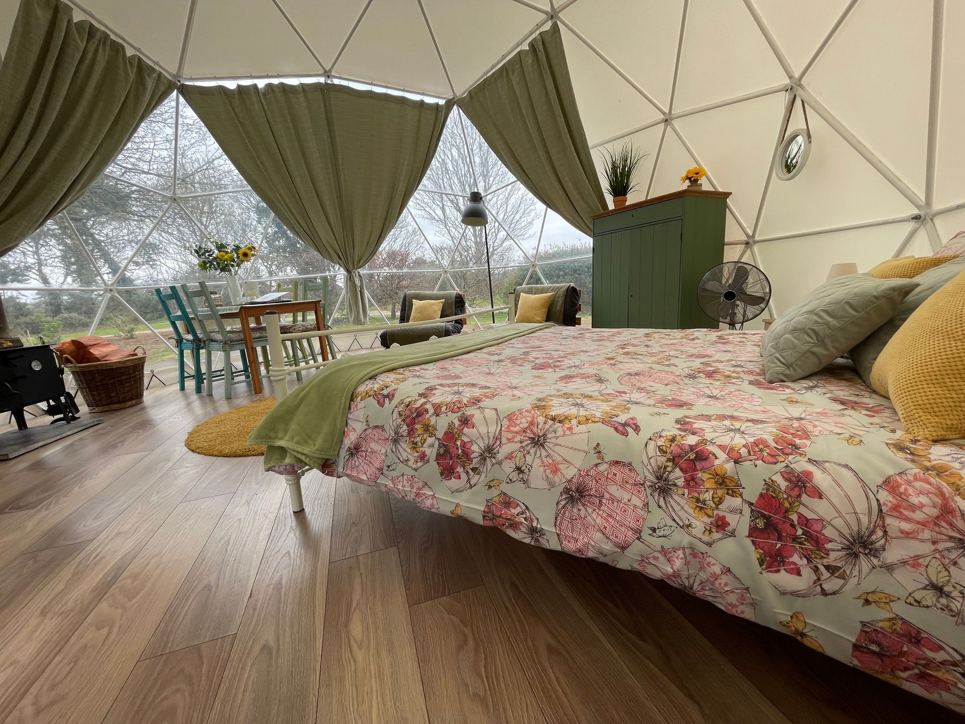 A cozy interior of a geodesic dome featuring a floral-print bed, green curtains, and a view of a wooded area outside.