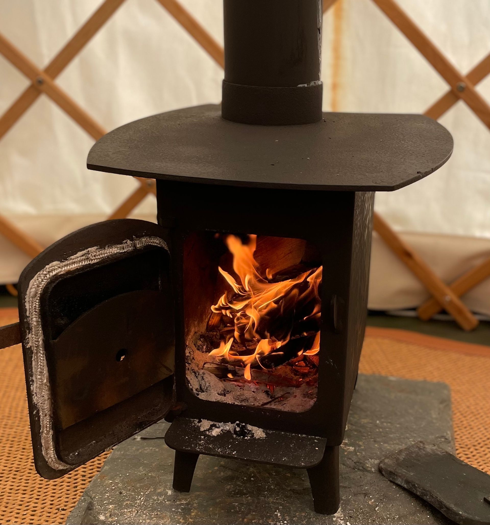A black wood-burning stove with a glowing fire inside, set on a stone base inside a white yurt with wooden lattice walls.