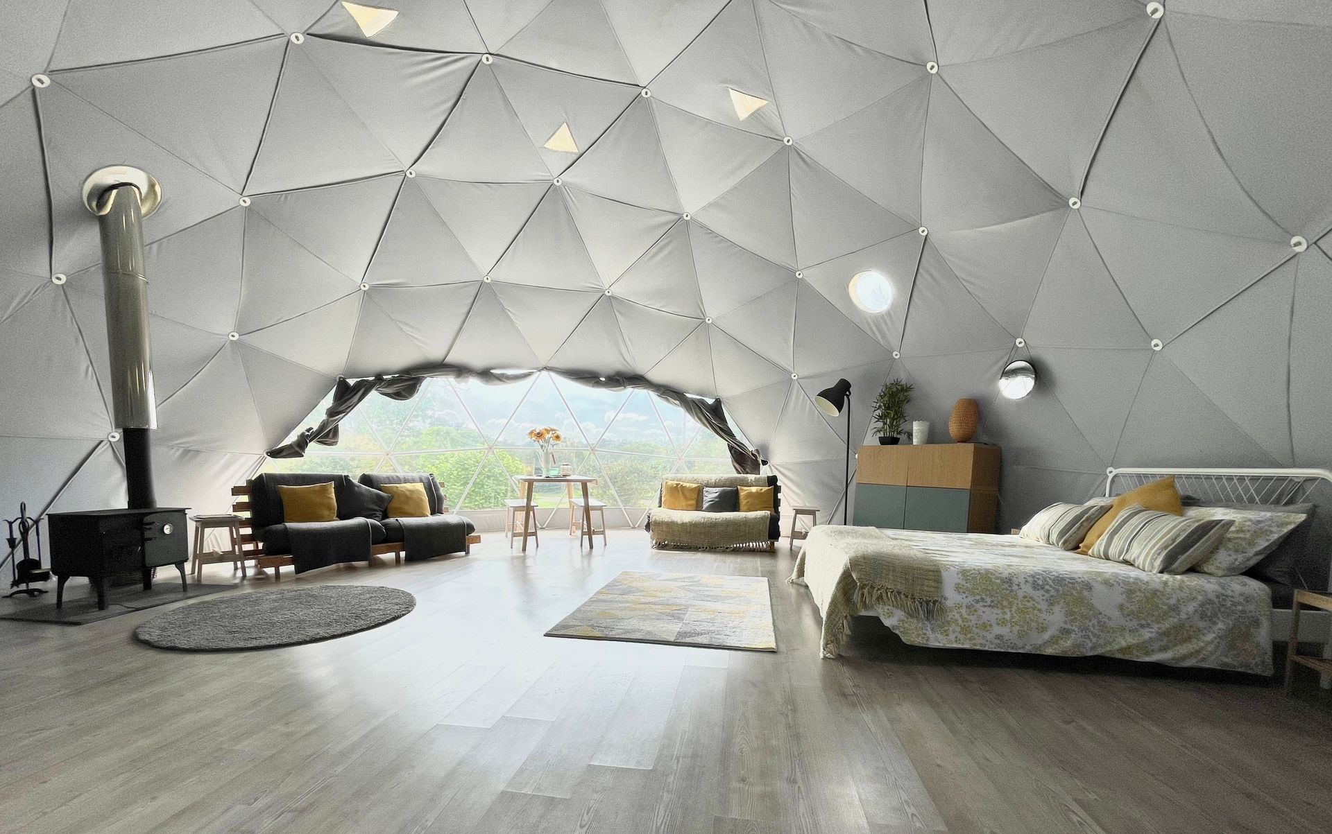Interior of a geodesic dome tent featuring light-colored triangular panels, a wood stove, seating, and a bed.