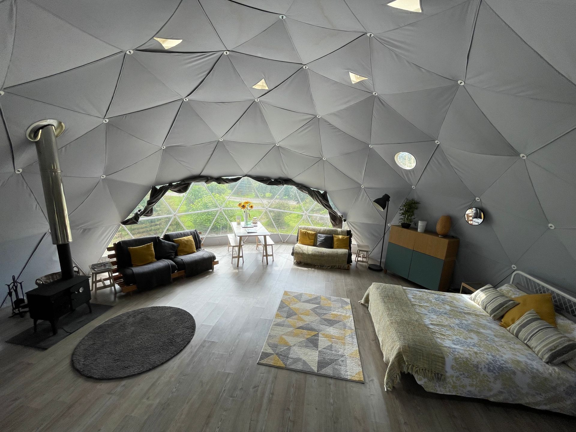 Interior of a spacious geodesic dome tent with a wood-burning stove, living area, and bed, featuring a large window.