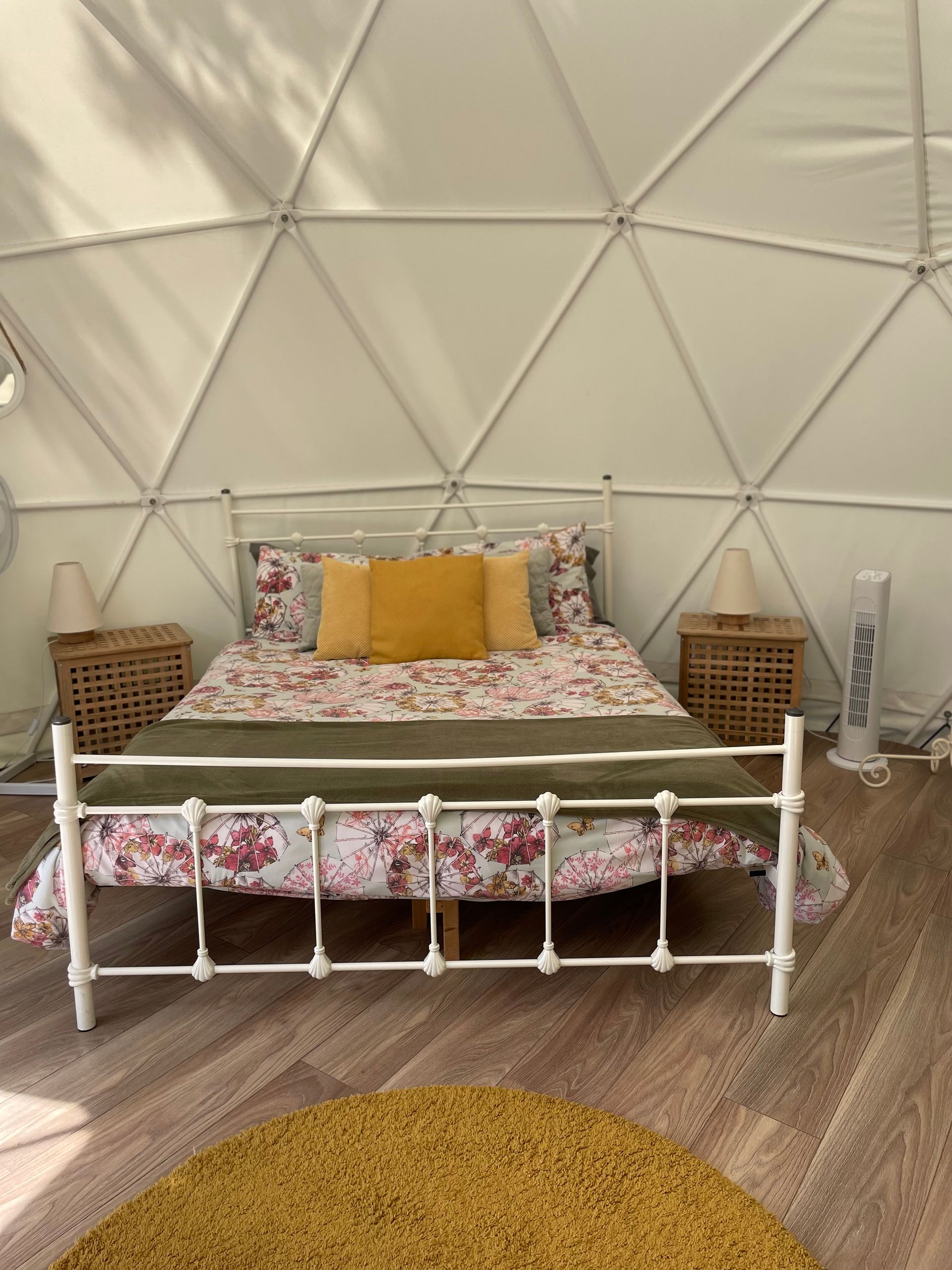 A white metal bed with floral bedding and yellow cushions inside a geodesic dome tent with wooden flooring.