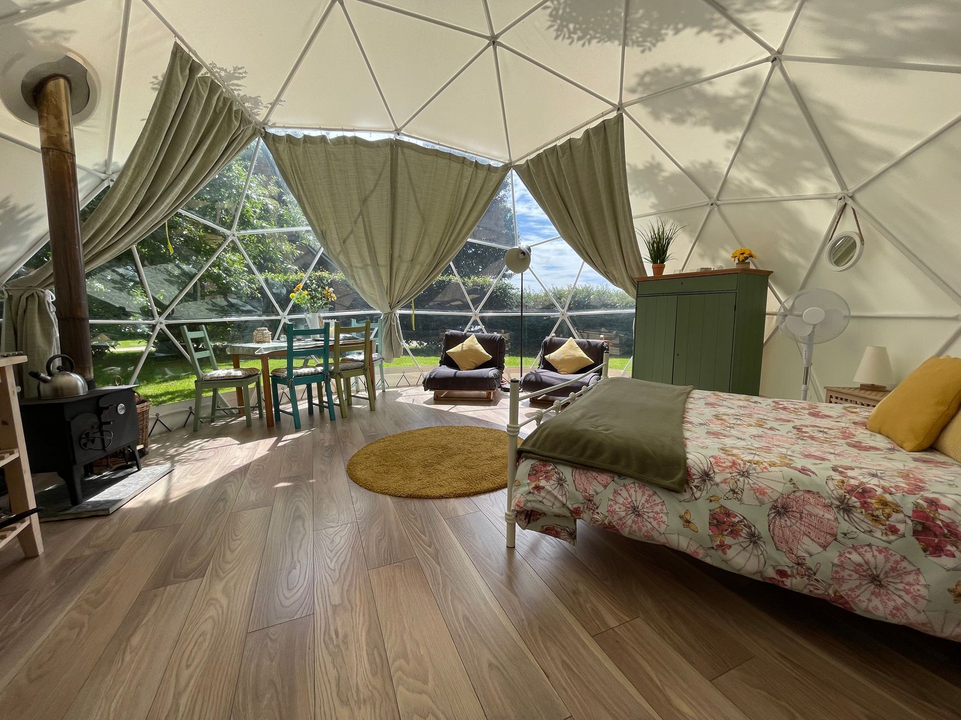 A cozy geodome interior with a bed, wood-burning stove, dining set, and floor-to-ceiling windows overlooking green trees.