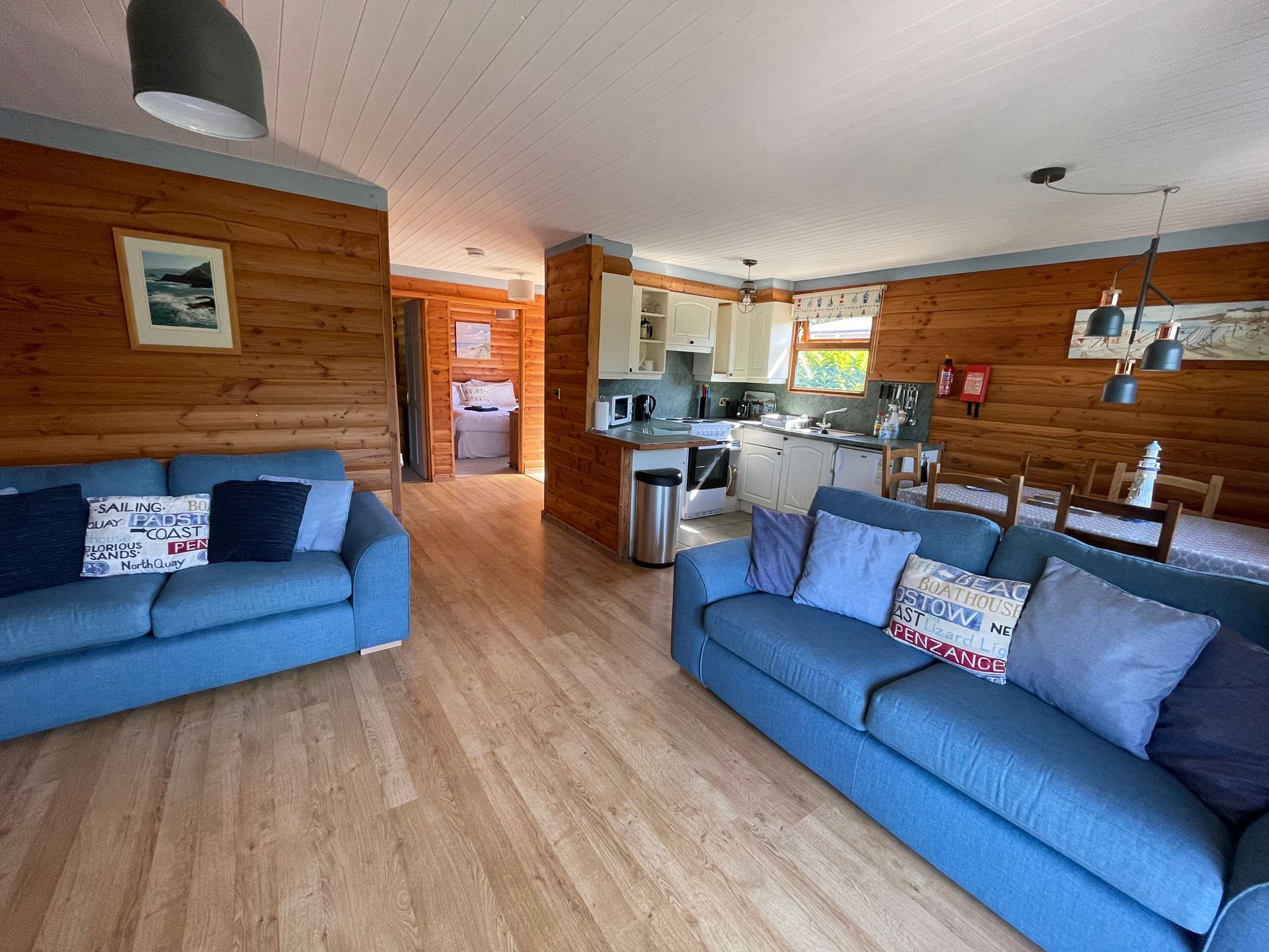 A spacious cabin living room featuring two blue sofas facing each other, wooden walls, and an open-plan kitchen behind.