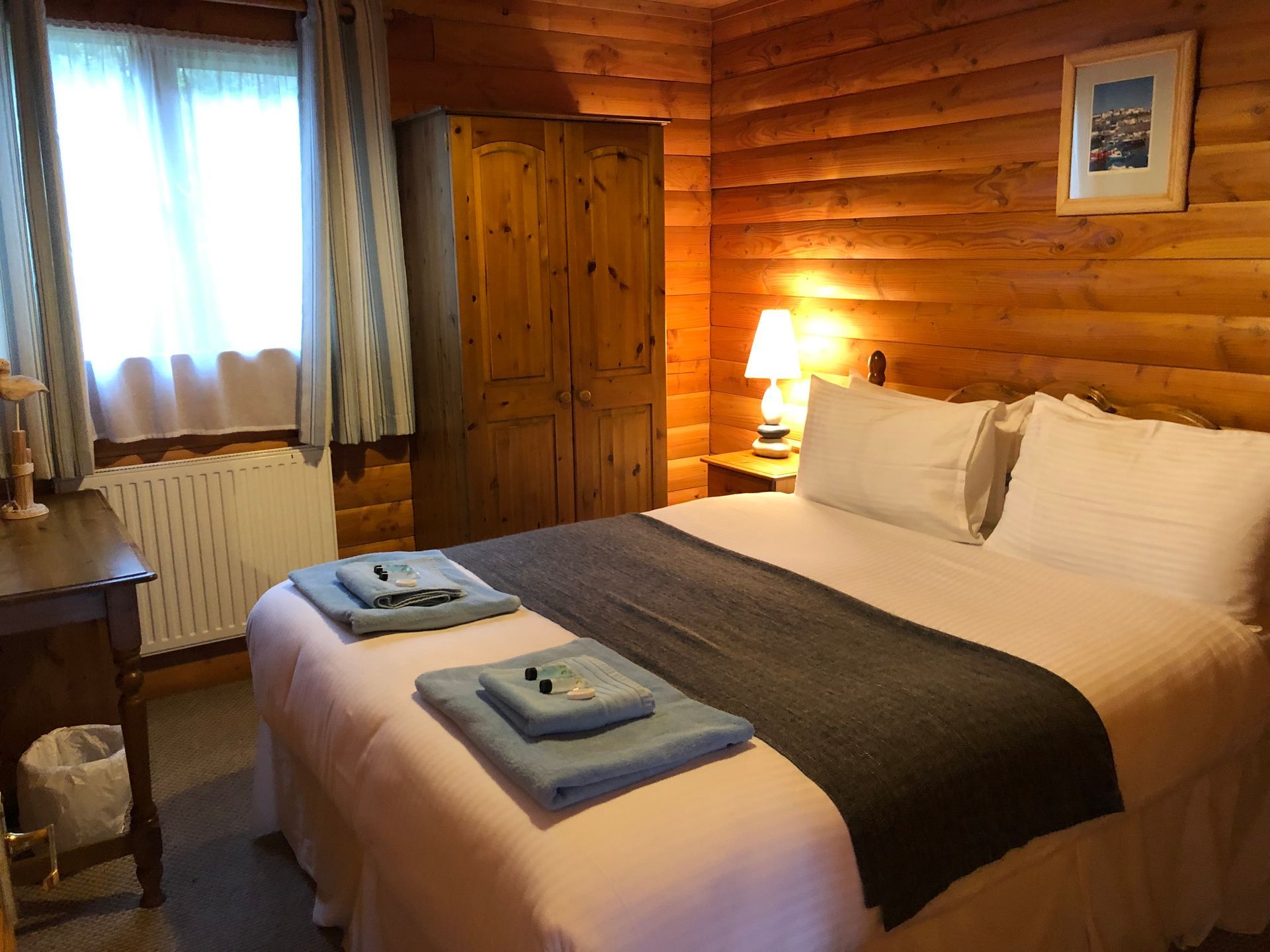 A cozy bedroom in a log cabin with a double bed, wooden walls, a wardrobe, and a window with curtains.