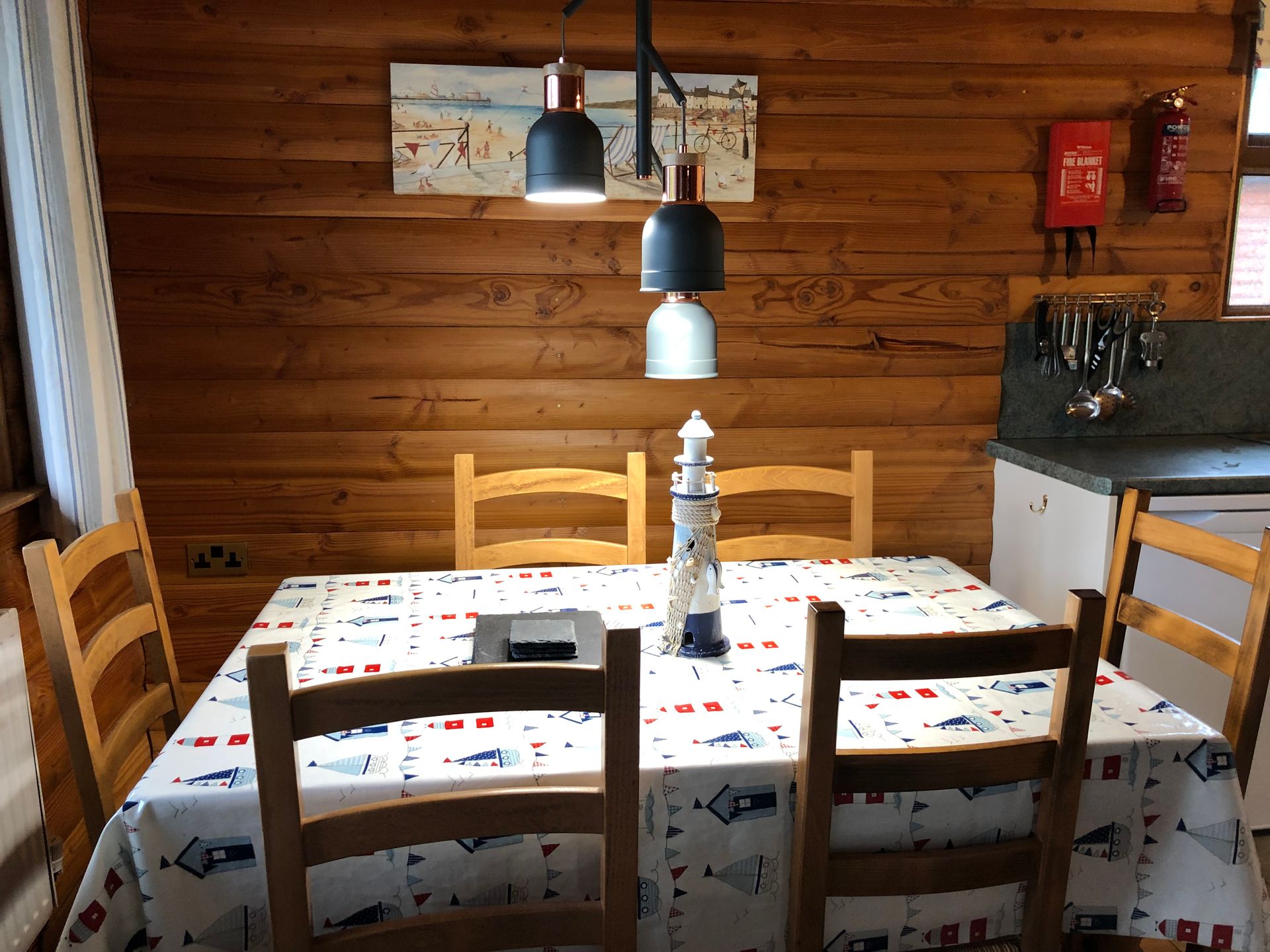 A wooden dining table with a nautical-themed tablecloth, chairs, and a lighthouse centerpiece under hanging lights.