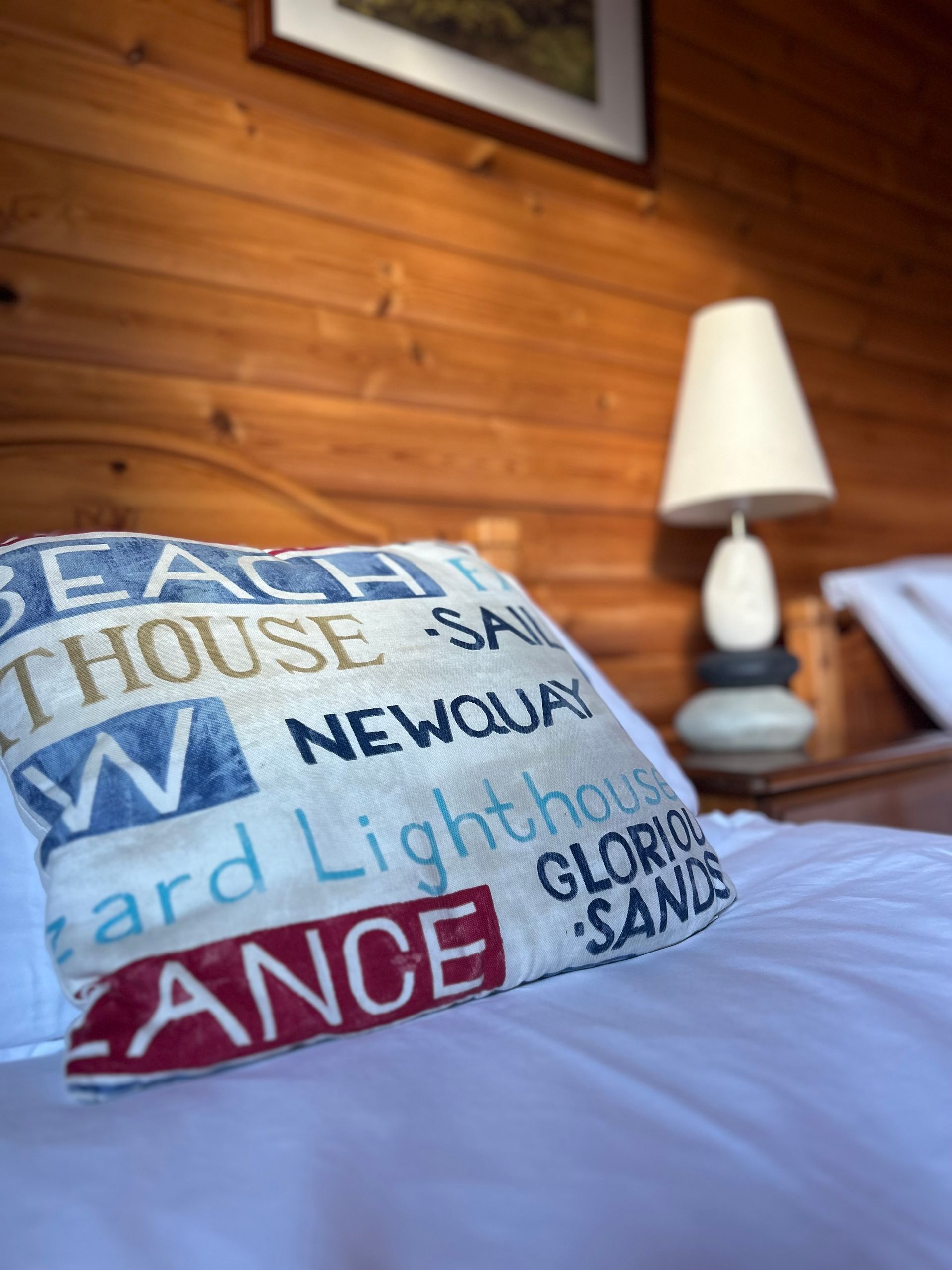 A throw pillow with coastal-themed text sits on a white bed in a room with warm, wooden-paneled walls and a bedside lamp.