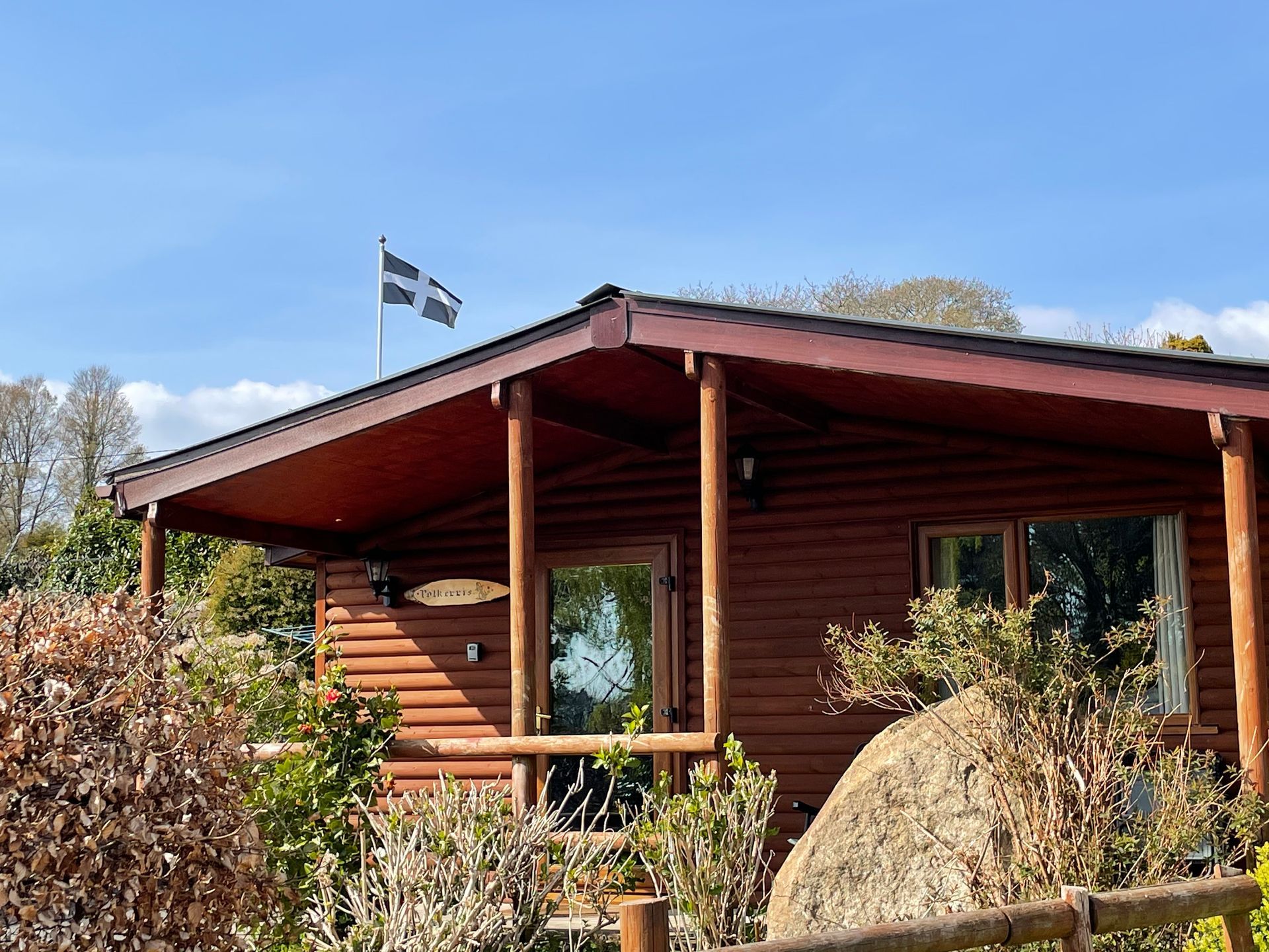 A wooden cabin with a porch under a clear blue sky, featuring a Saint Piran's flag flying from the roof.
