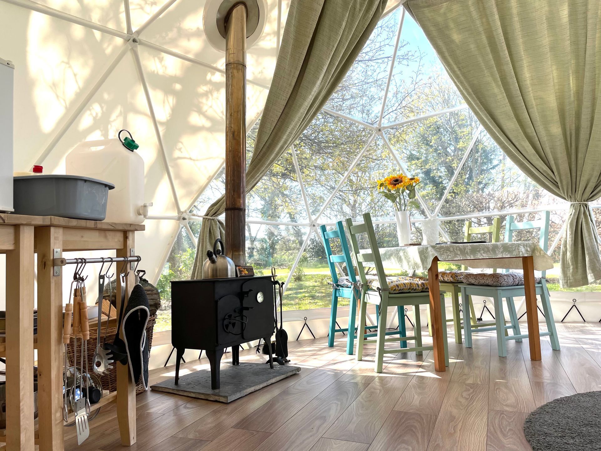 Interior of a geodesic dome tent with a wood-burning stove, wooden dining table with colorful chairs, and green curtains.