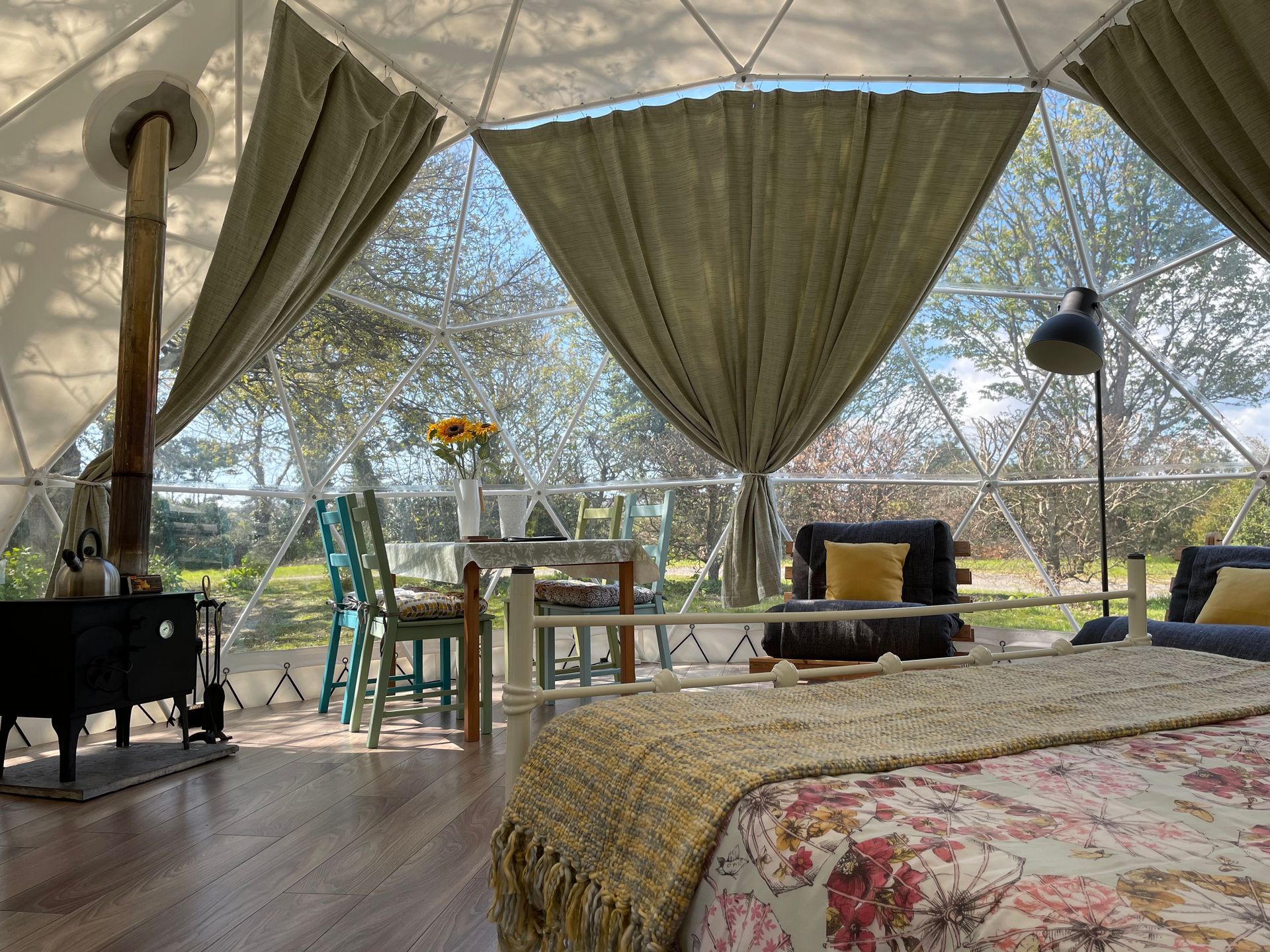 A cozy, light-filled geodesic dome glamping tent featuring a bed, a dining table with chairs, and a black wood-burning stove.
