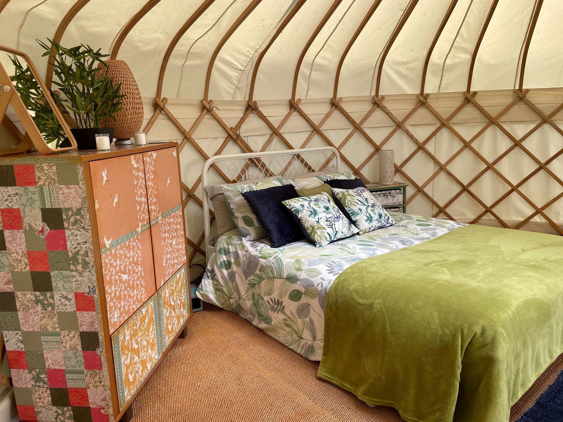 A cozy yurt interior featuring a bed with patterned linens, a green throw blanket, and a colorful patterned dresser.