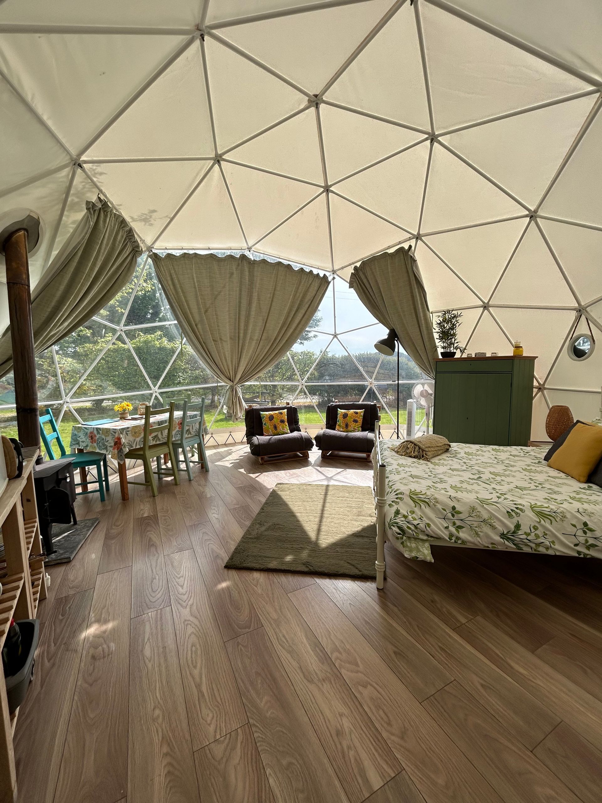 Interior of a bright, spacious geodesic dome tent with wooden floors, a bed, a dining area, and large draped windows.