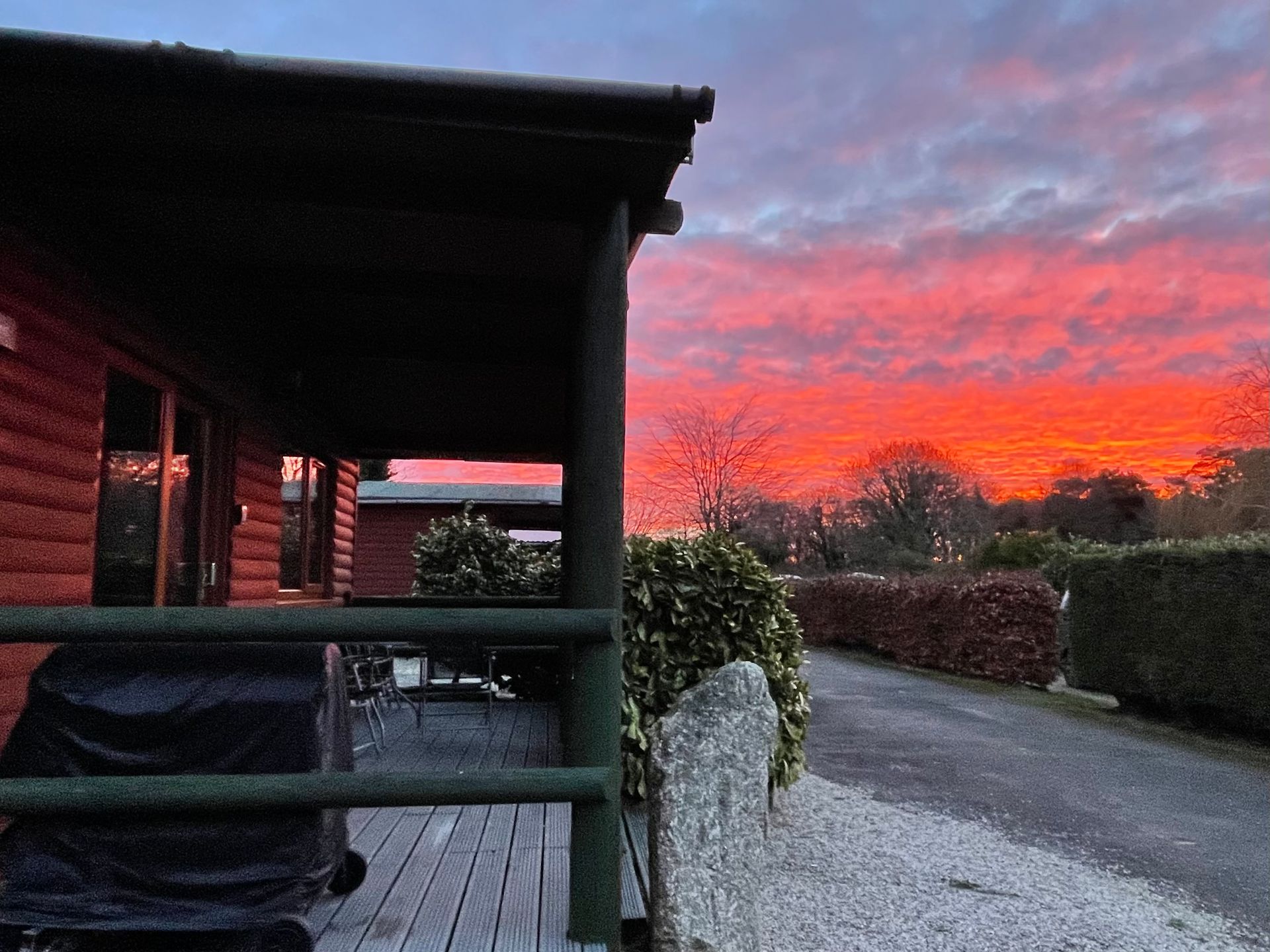 A red log cabin with a porch overlooks a gravel path and hedge beneath a vibrant, fiery orange and purple sunset.
