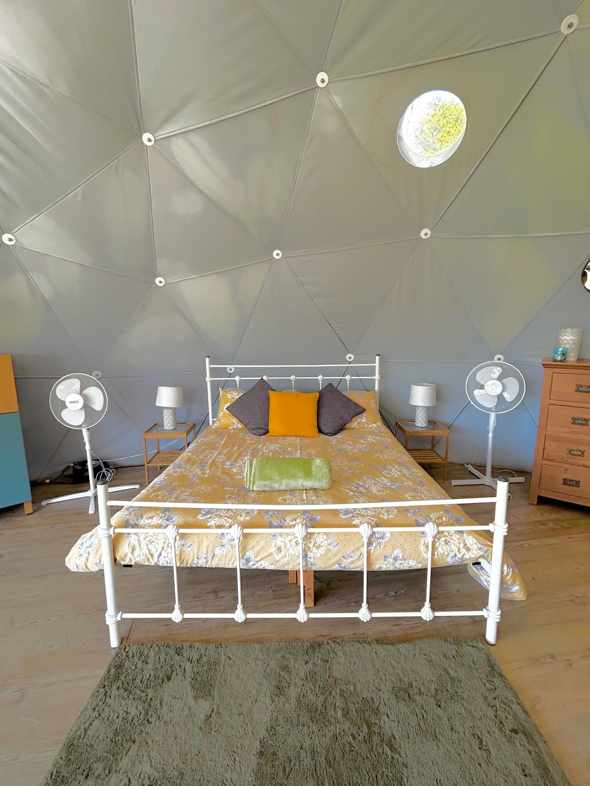 A bed with yellow patterned bedding, two fans, and nightstands inside a geometric dome tent structure.