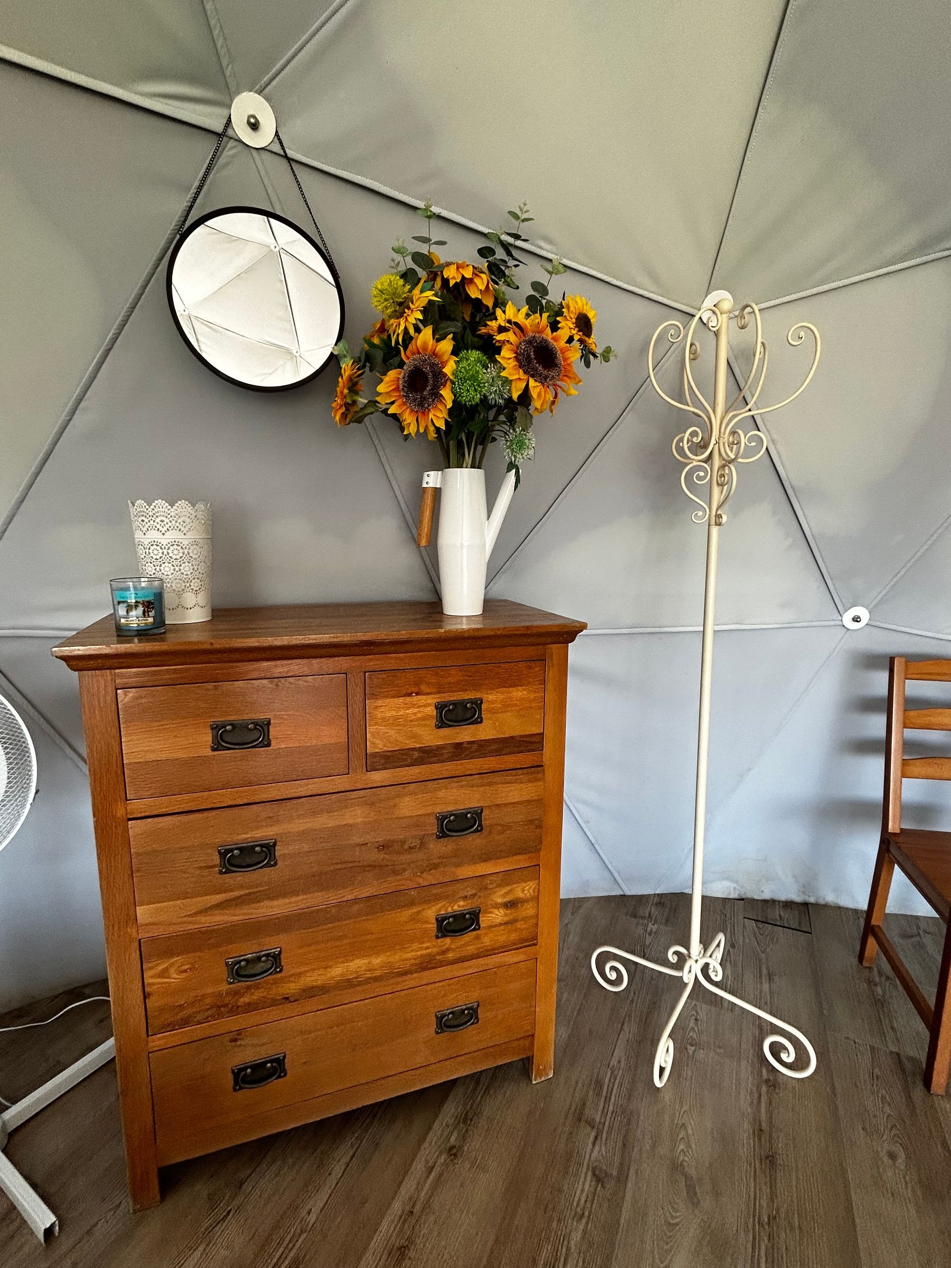 A wooden dresser holding a flower vase, a candle, and a small lantern, next to a metal coat rack and mirror in a dome tent.