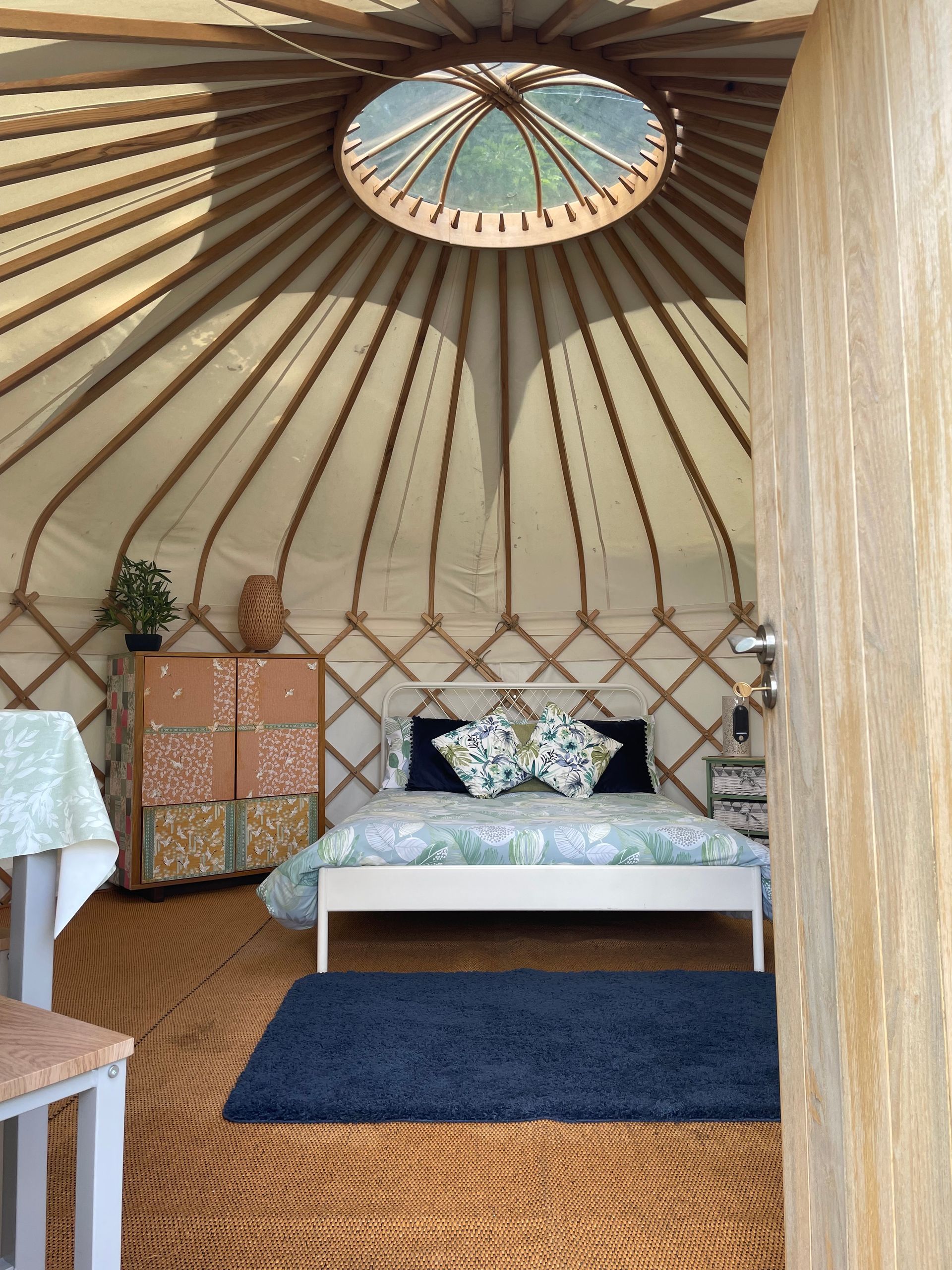 Interior view of a yurt with a central circular skylight, a bed with patterned pillows, a patterned cabinet, and a rug.