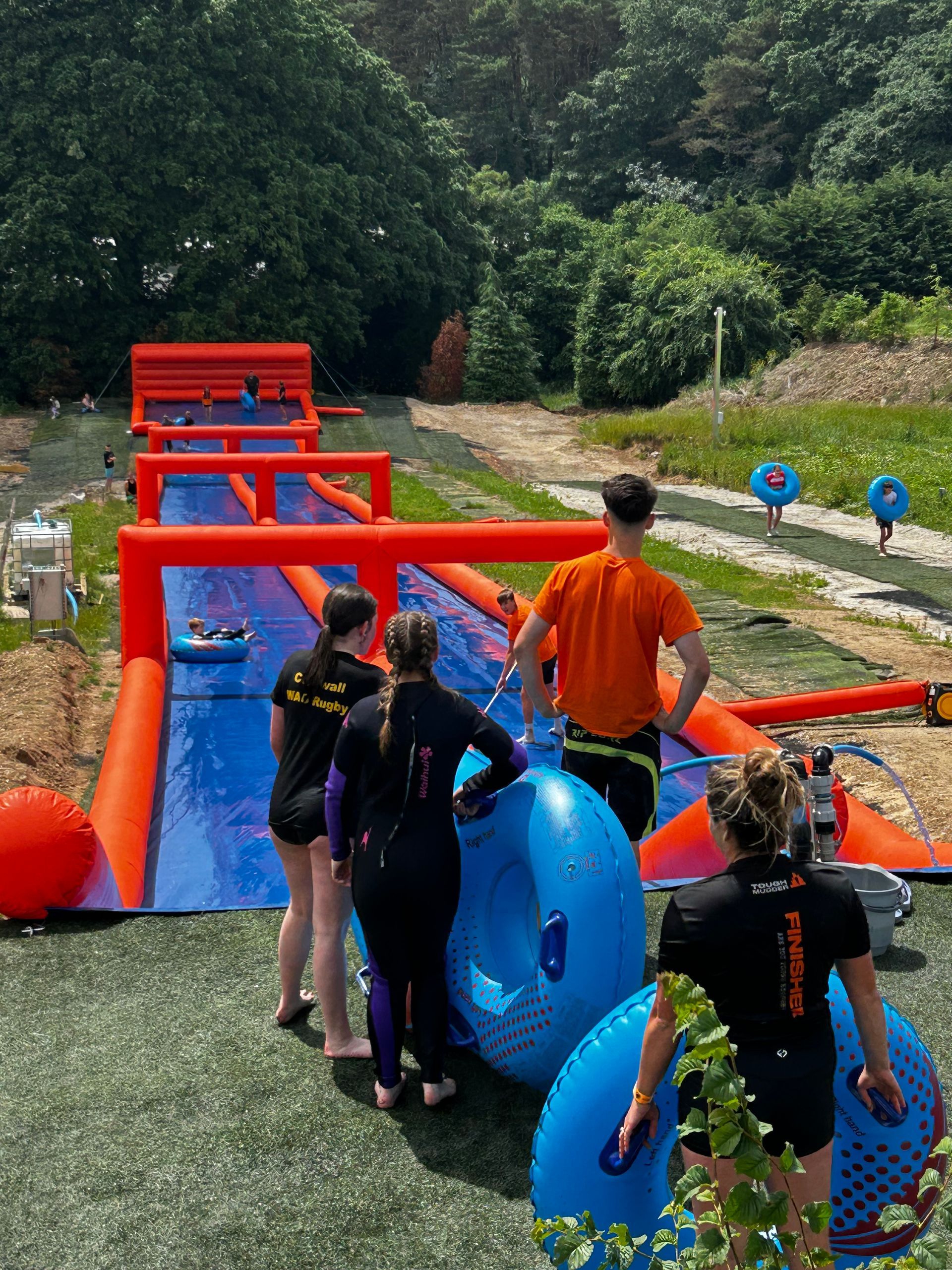 People holding blue tubes wait at the top of a long, red inflatable water slide at an outdoor park.