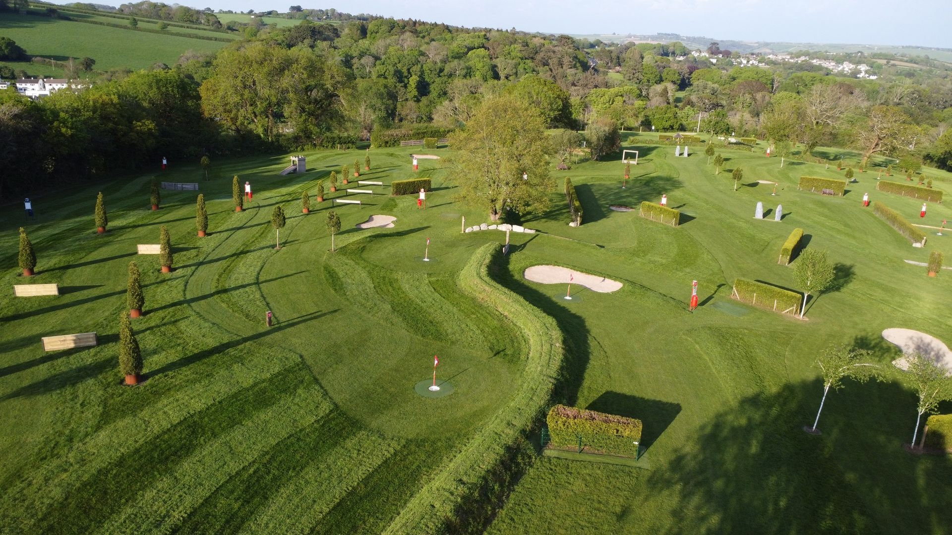 Aerial view of a grass cross-country equestrian course with various wooden jumps and hedges scattered across rolling hills.
