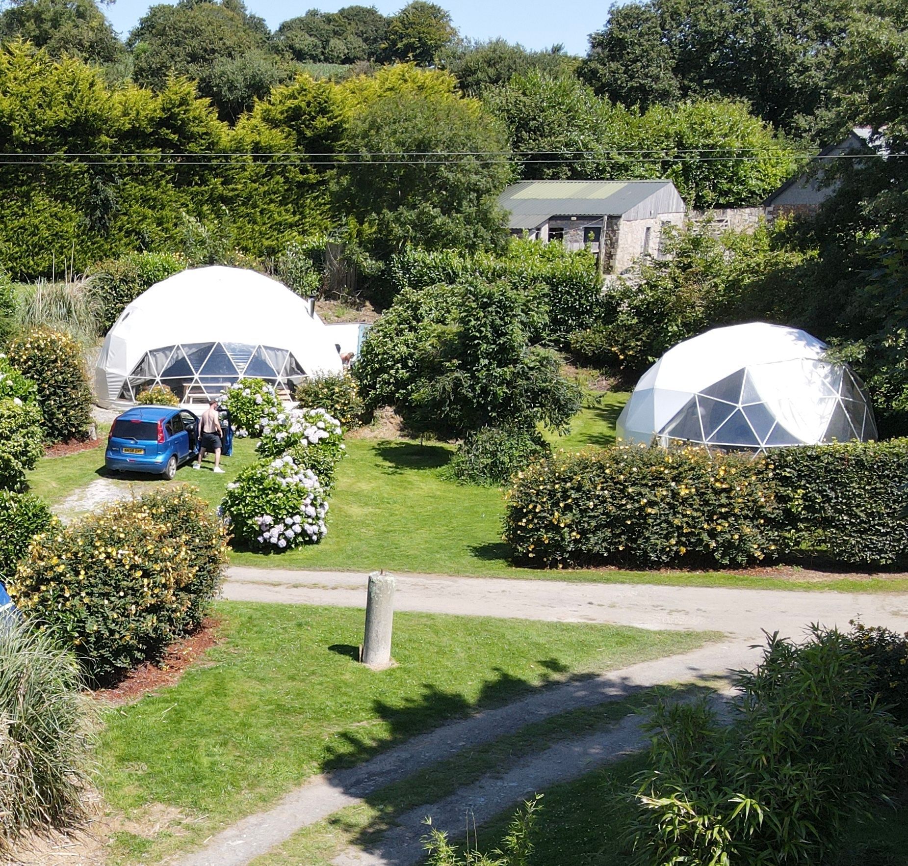 Two white geodesic domes sit in a lush green garden with a blue car parked beside one and a gravel path in the foreground.