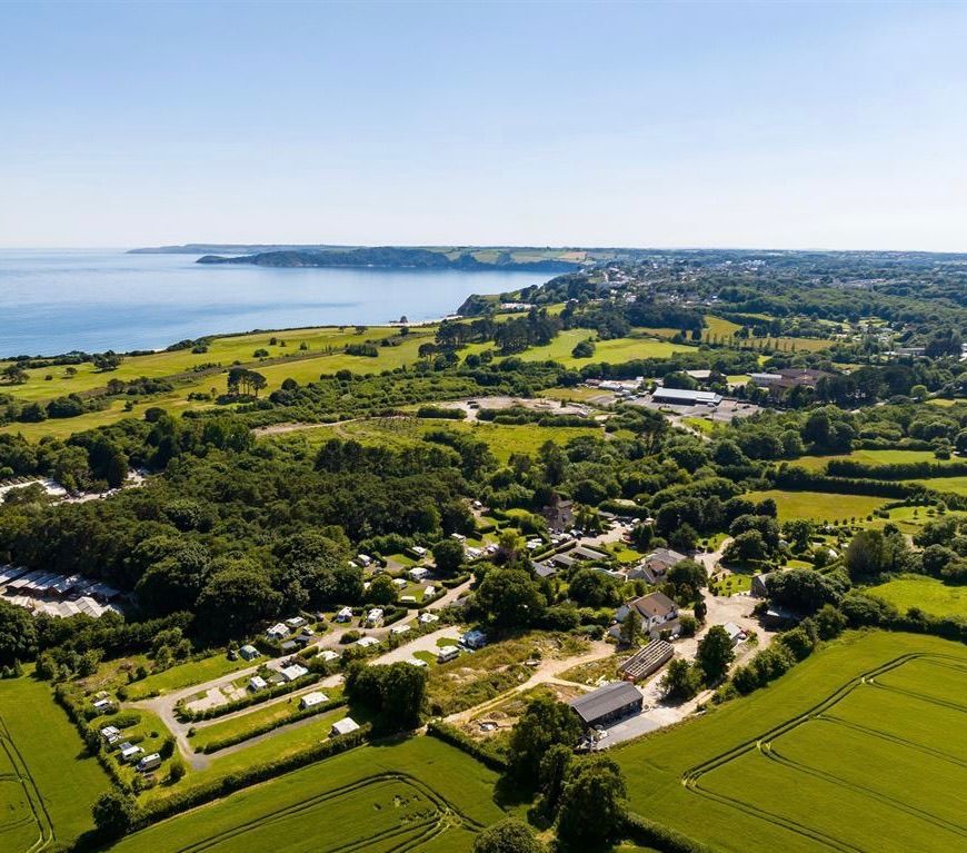 Aerial view of a coastal campsite nestled in lush green fields and trees, overlooking a calm blue sea under a clear sky.
