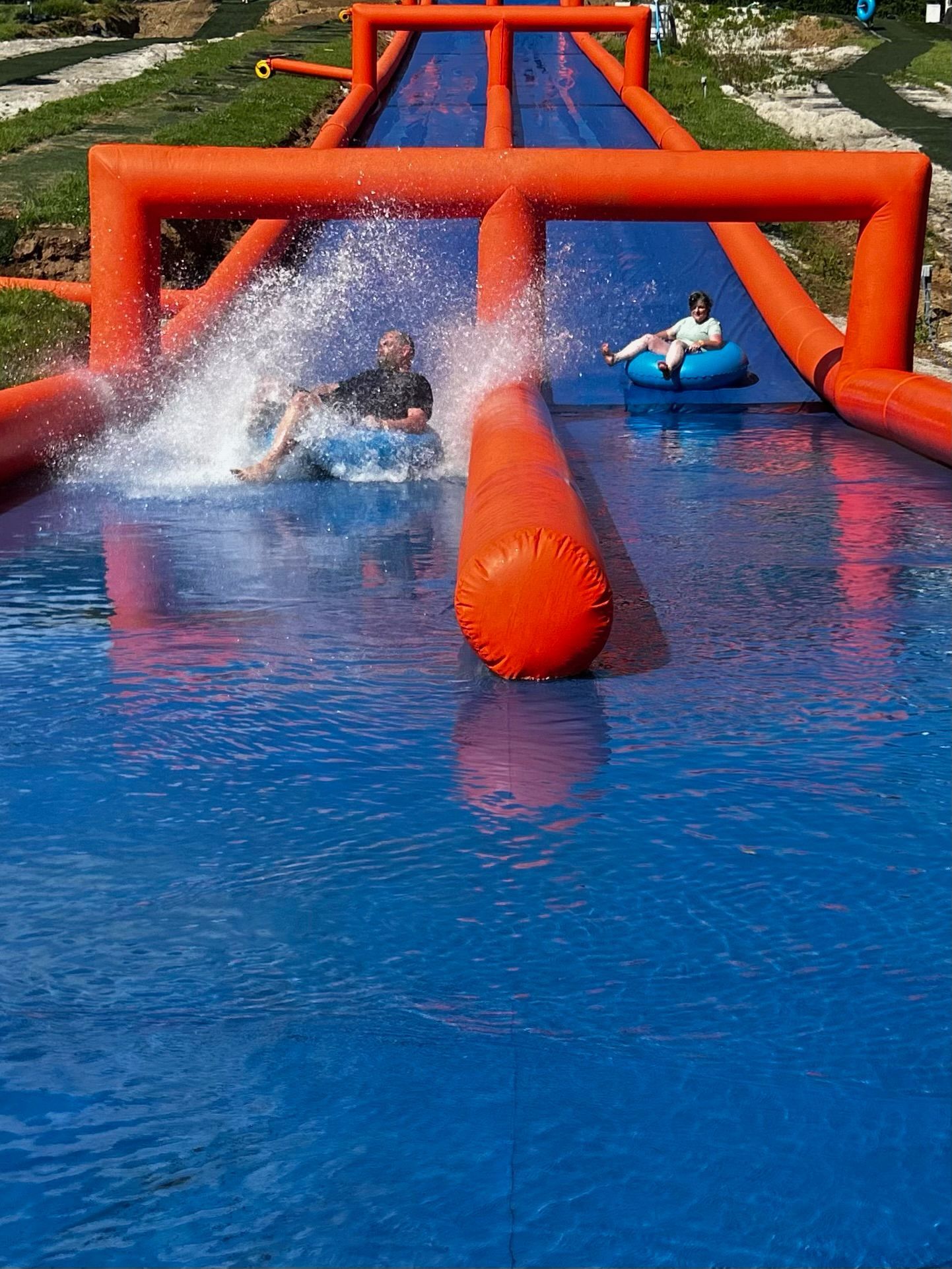 Two people slide down a blue water slide in inflatable tubes through a bright orange frame structure.