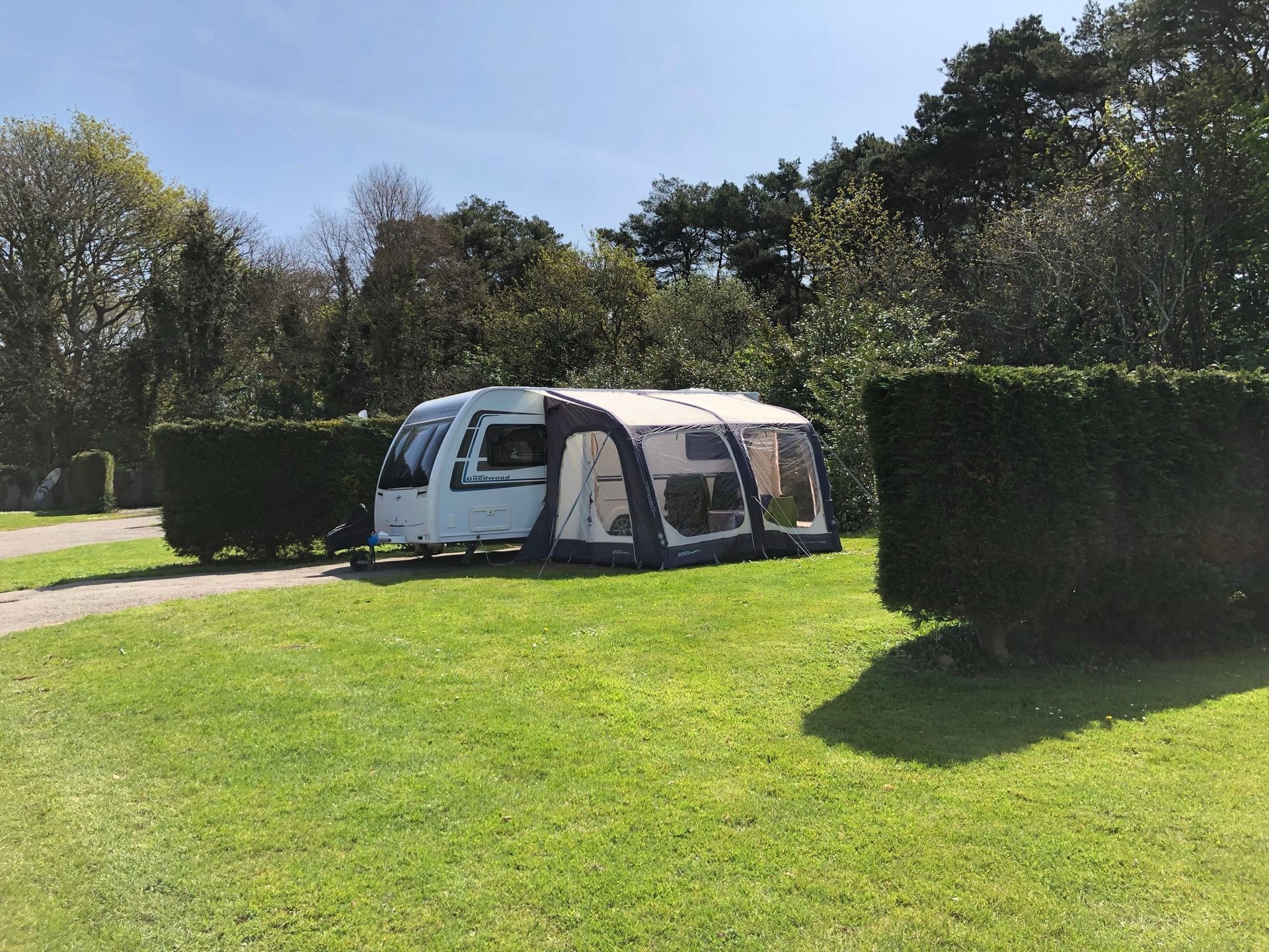 A white travel trailer with an attached grey inflatable awning, parked on a grassy site between two tall green hedges.