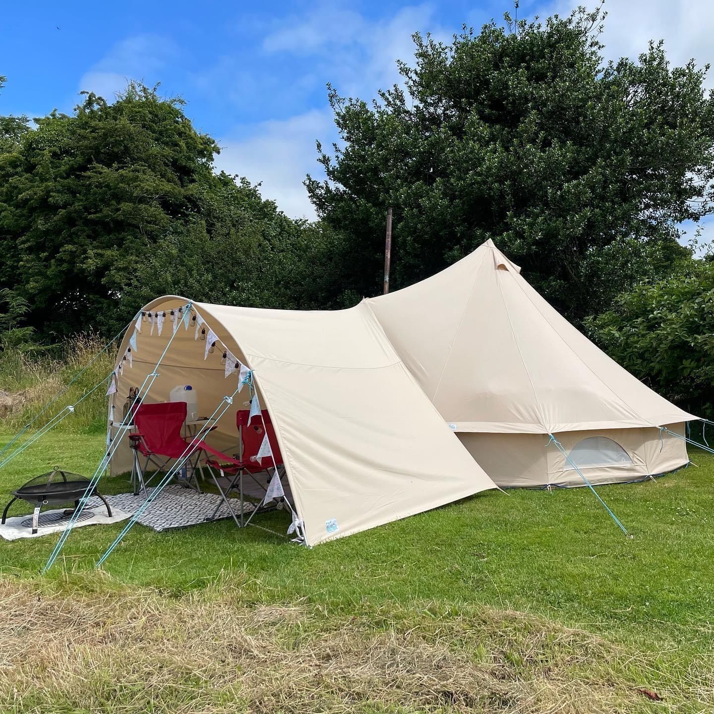 A beige bell tent with an attached canopy awning, set up on a green lawn surrounded by trees.