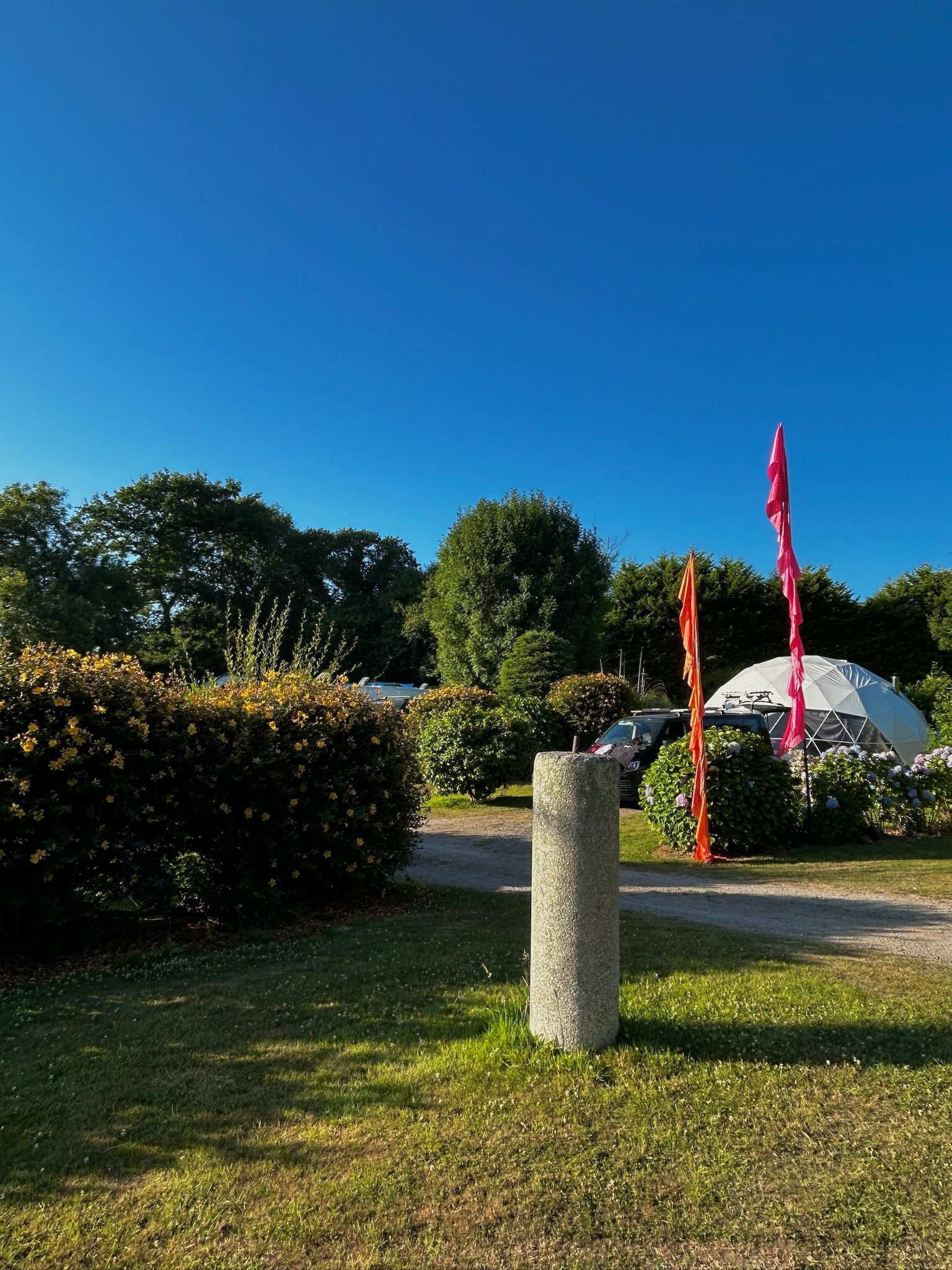 A grassy campsite with a stone pillar in the foreground, bushes, trees, and a white dome tent under a bright blue sky.