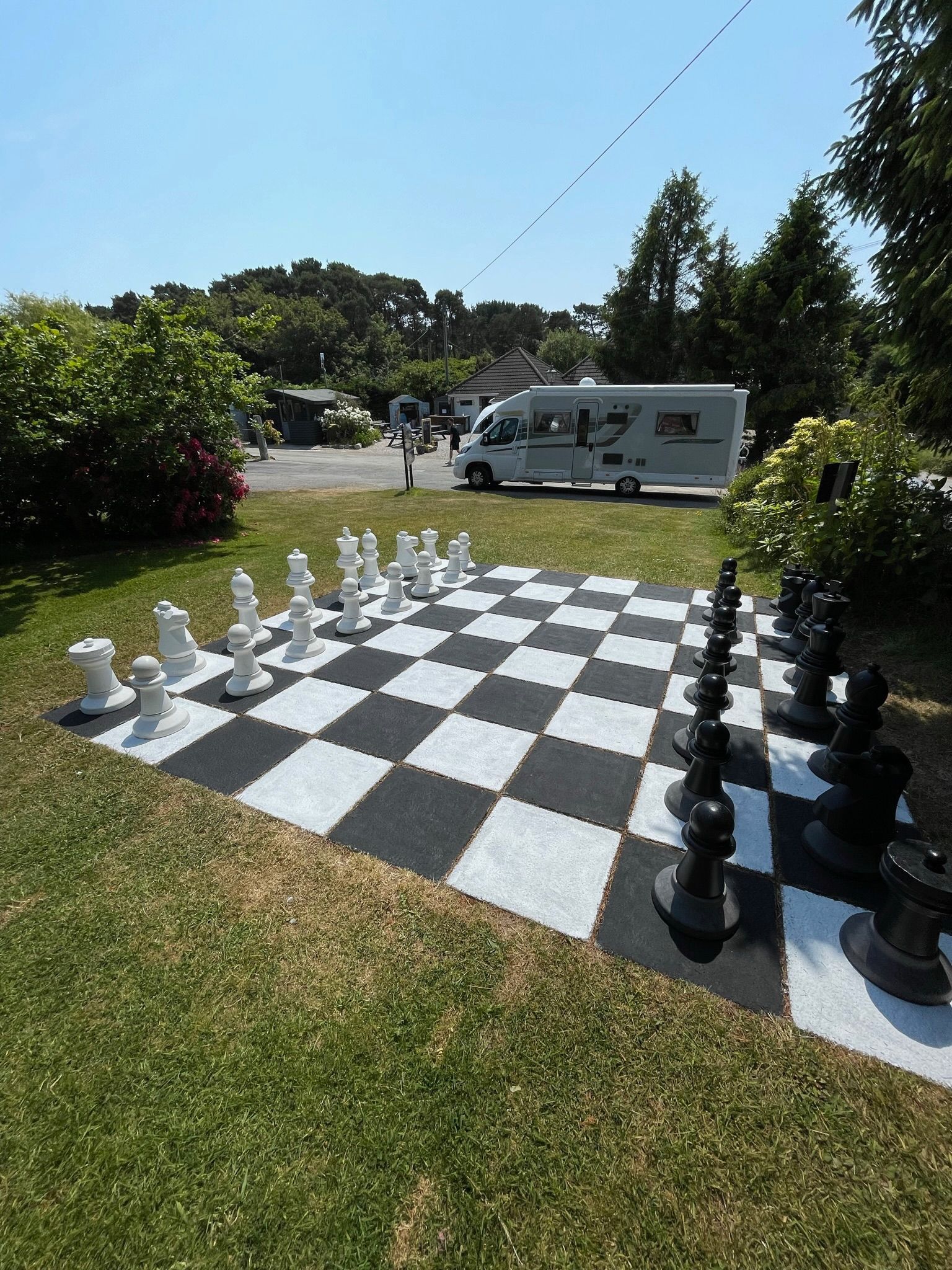 A giant outdoor chess set with black and white pieces on a checkered mat in a grassy area with a camper van nearby.
