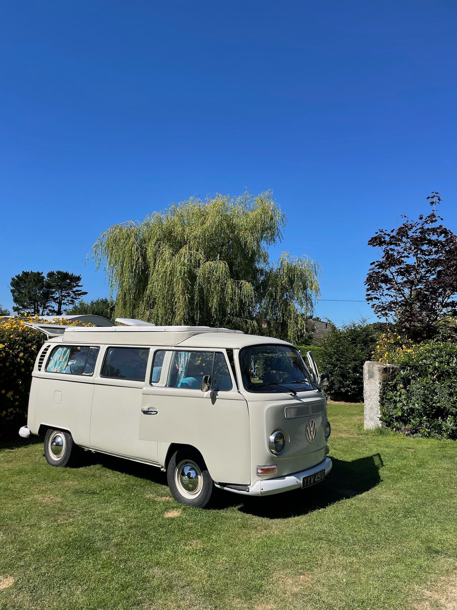 A beige vintage Volkswagen camper van parked on a grassy lawn under a clear blue sky.