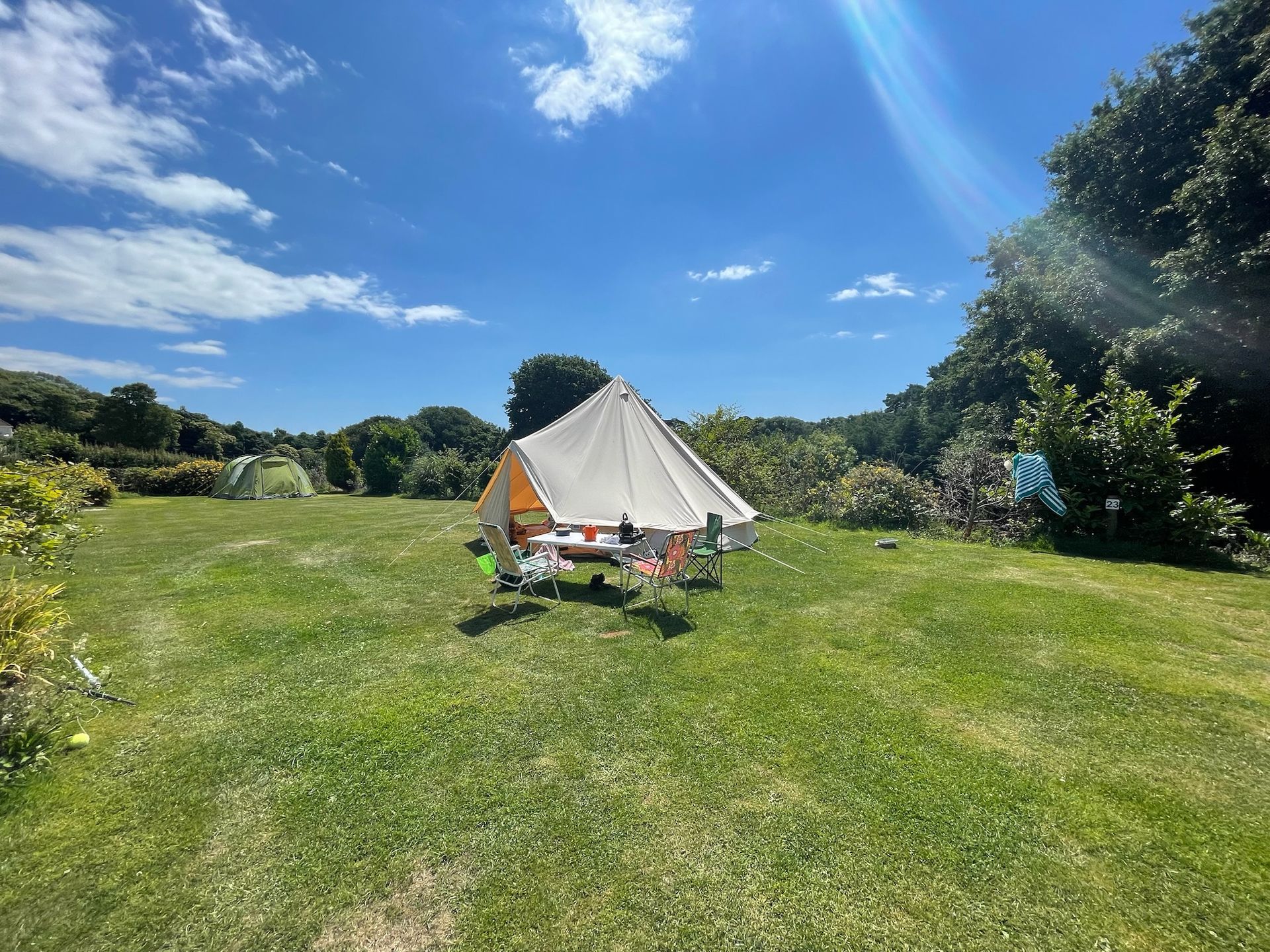 A canvas bell tent sits in a sunlit grassy field under a bright blue sky, surrounded by trees and shrubs.