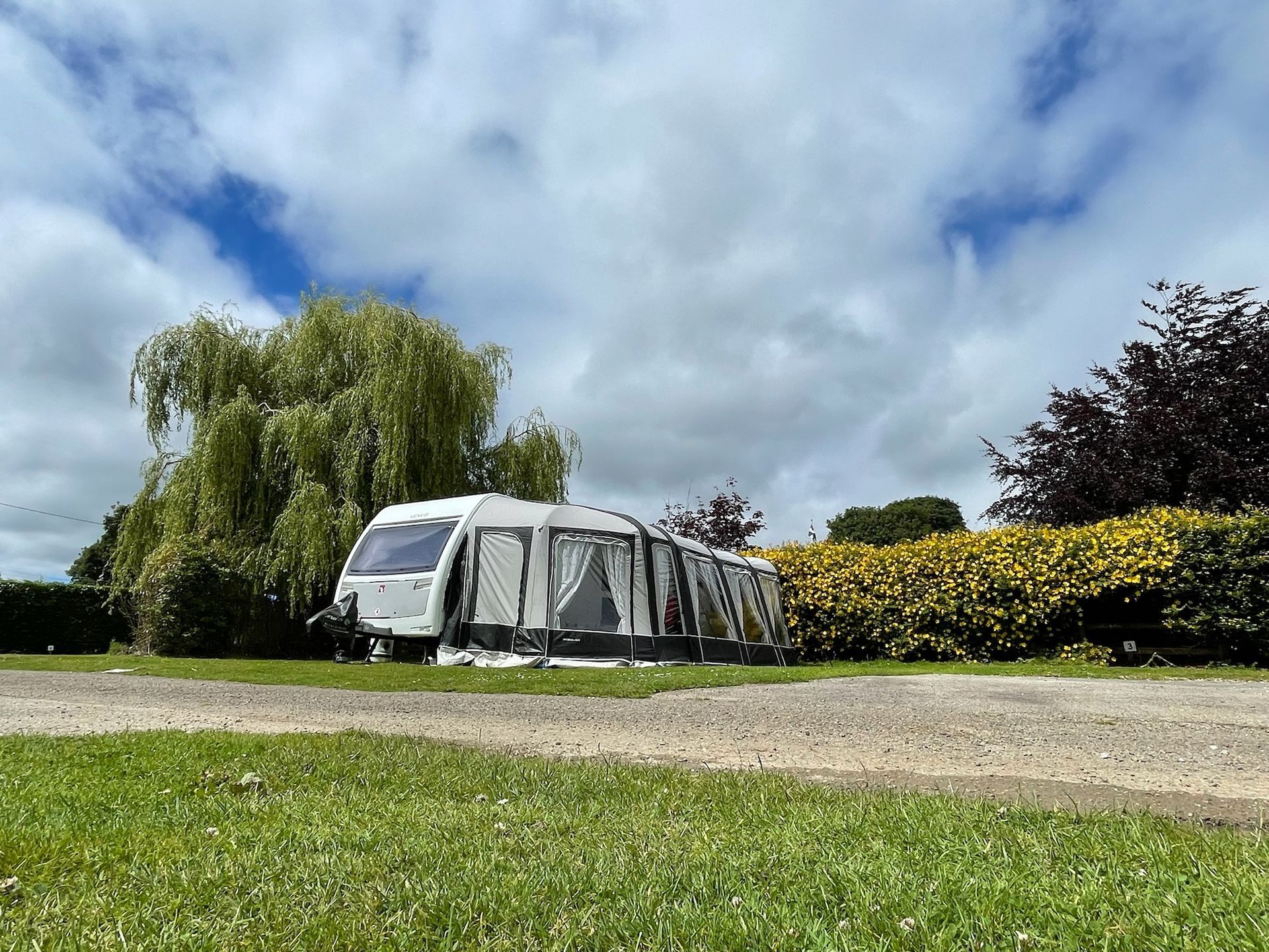 A white caravan with an attached gray awning parked on grass next to a willow tree and a hedge under a cloudy blue sky.