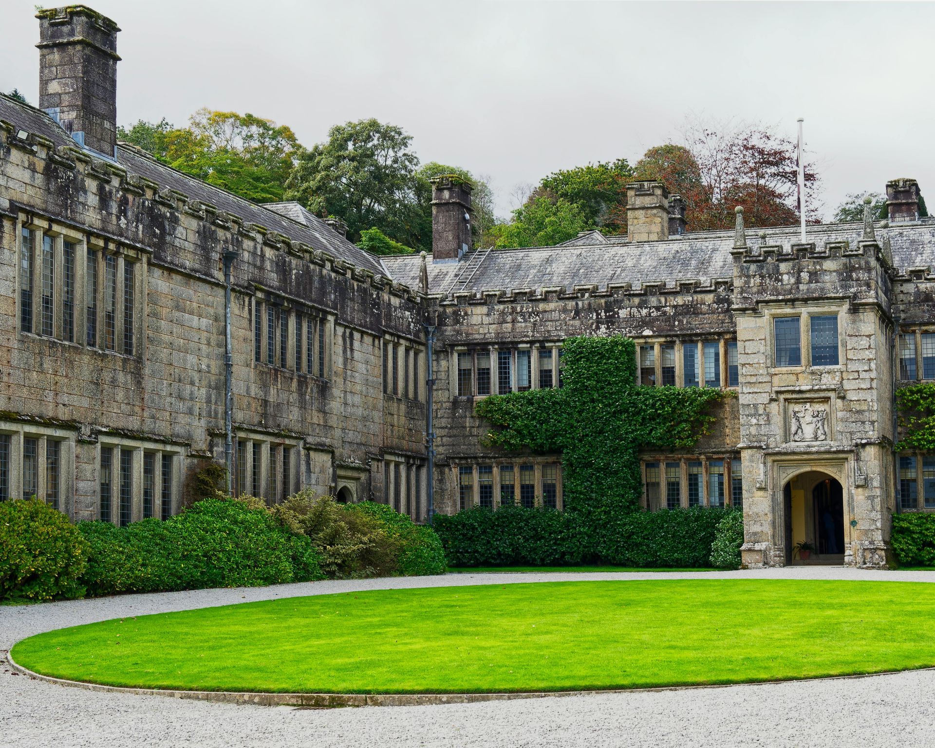An old stone manor house featuring a central arched entryway and windows covered in ivy, set behind a circular lawn.
