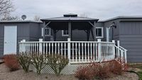 Front porch of a gray house with white railing, screened entry, and leafless shrubs in front