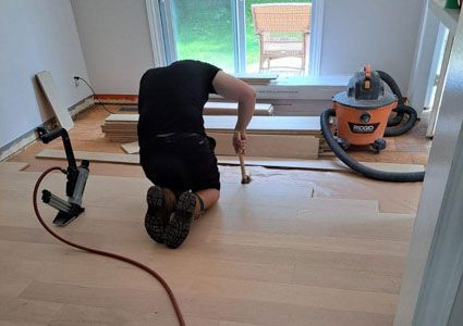 Person kneeling on a hardwood floor, sanding boards during a room renovation, with a vacuum nearby