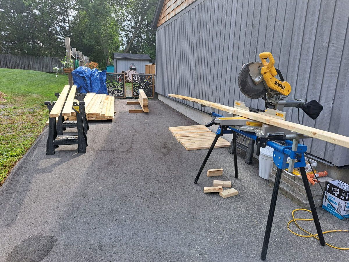 Outdoor work area with lumber on sawhorses, a miter saw, and tools beside a building.