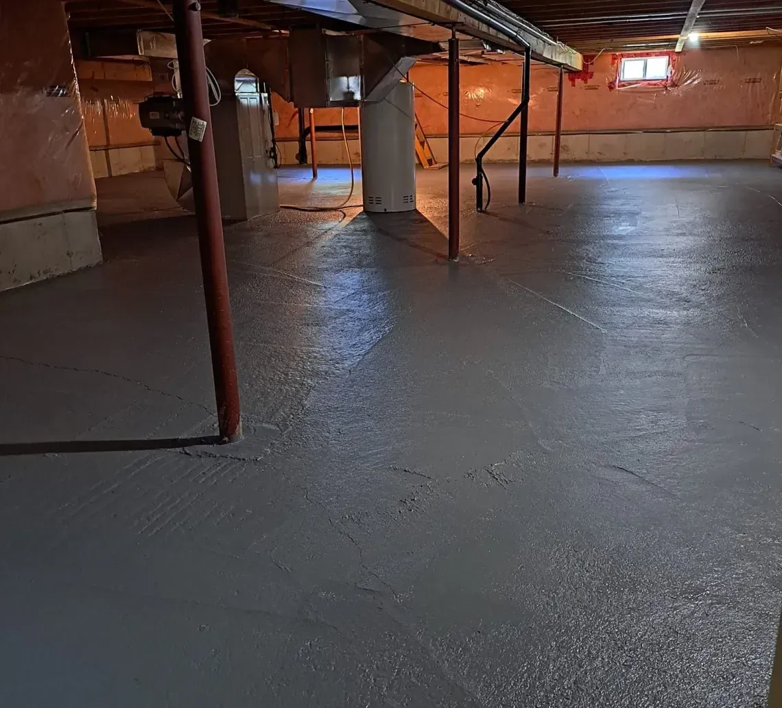 Basement crawlspace with low beams, support posts, and a wet, gray floor under warm lighting