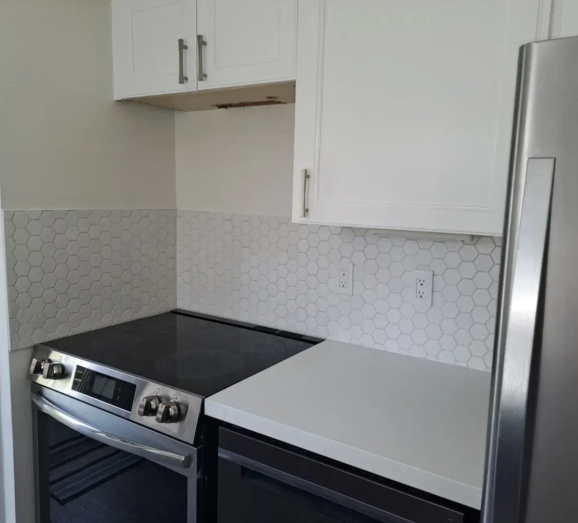 Kitchen corner with stainless steel stove and refrigerator, white cabinets, and speckled tile backsplash.