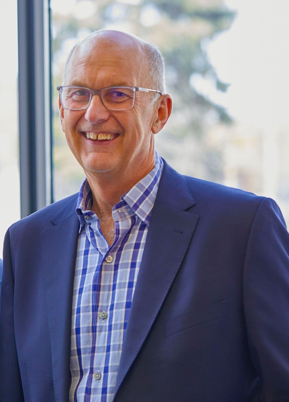 A man in a suit and plaid shirt is smiling in front of a window.