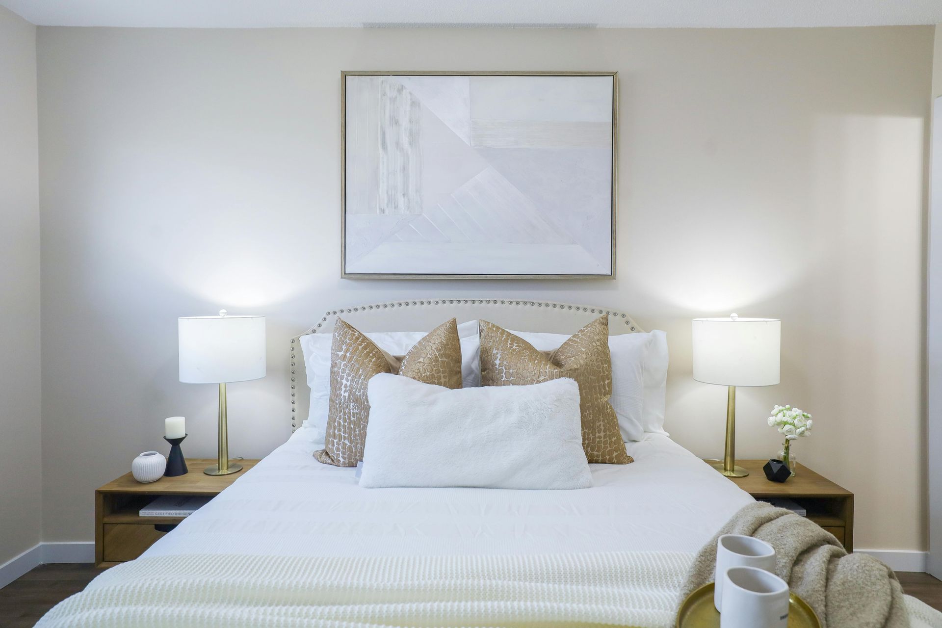 A bedroom featuring a bed with white bedding, gold patterned pillows, a central framed art print, and two bedside lamps.