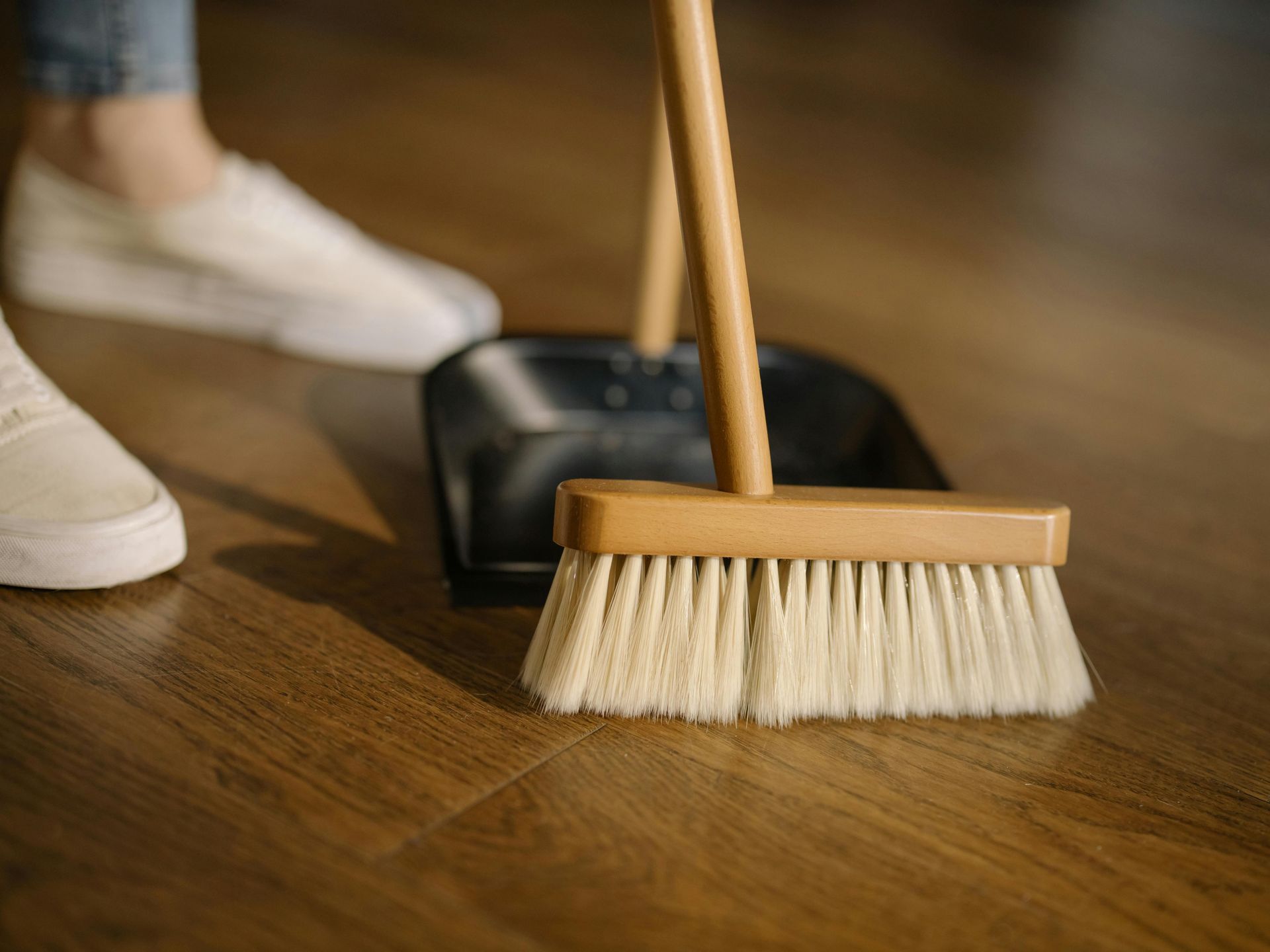 Close-up of a broom sweeping into a black dustpan on a wood floor, with feet in white sneakers visible nearby.
