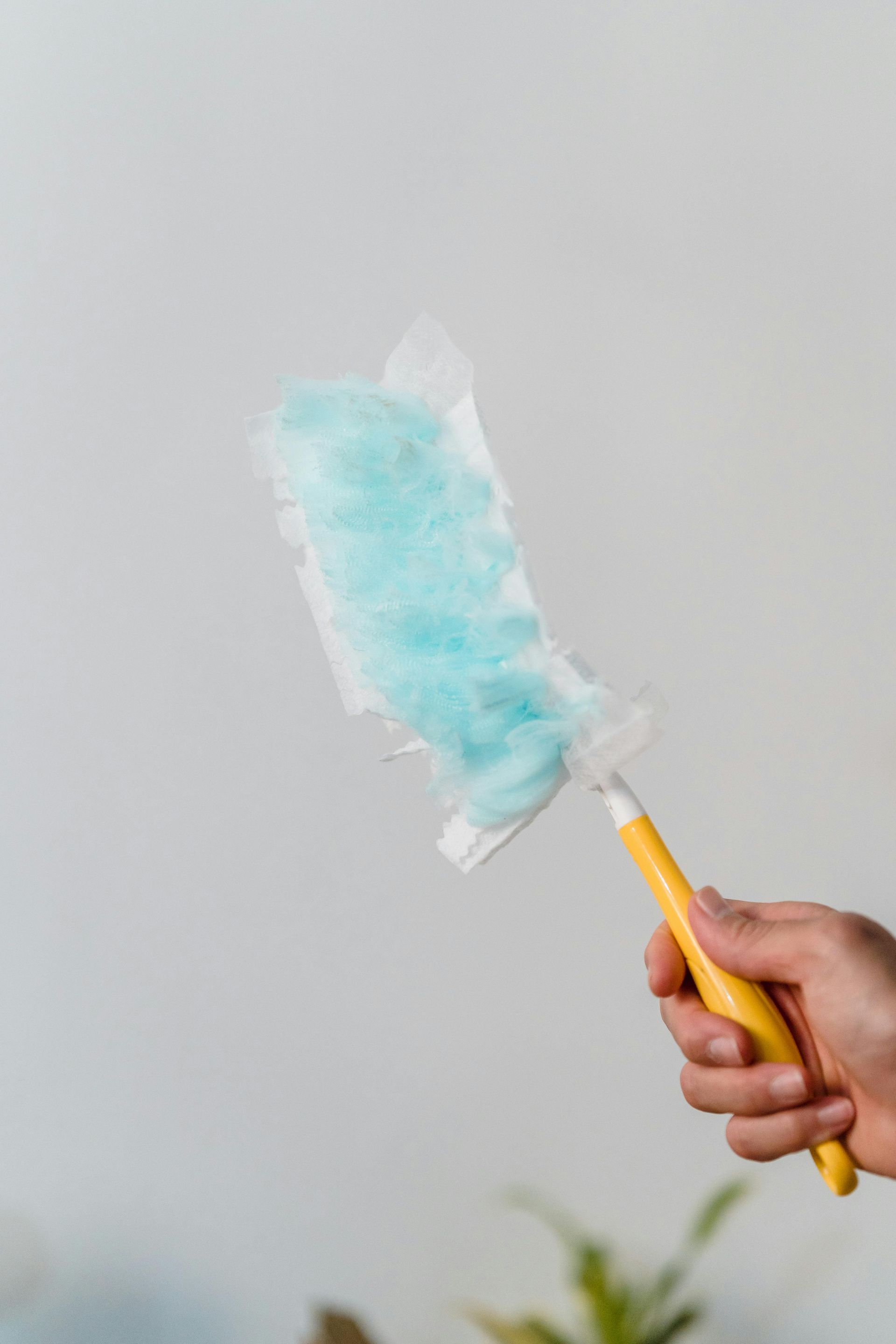 A hand holds a yellow-handled duster with a fluffy, light-blue disposable cleaning pad against a plain white background.