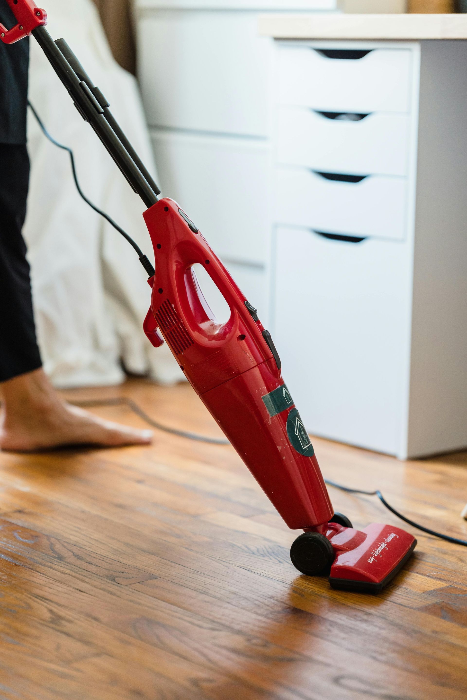 A person uses a red stick vacuum to clean a wooden floor near a white drawer unit.
