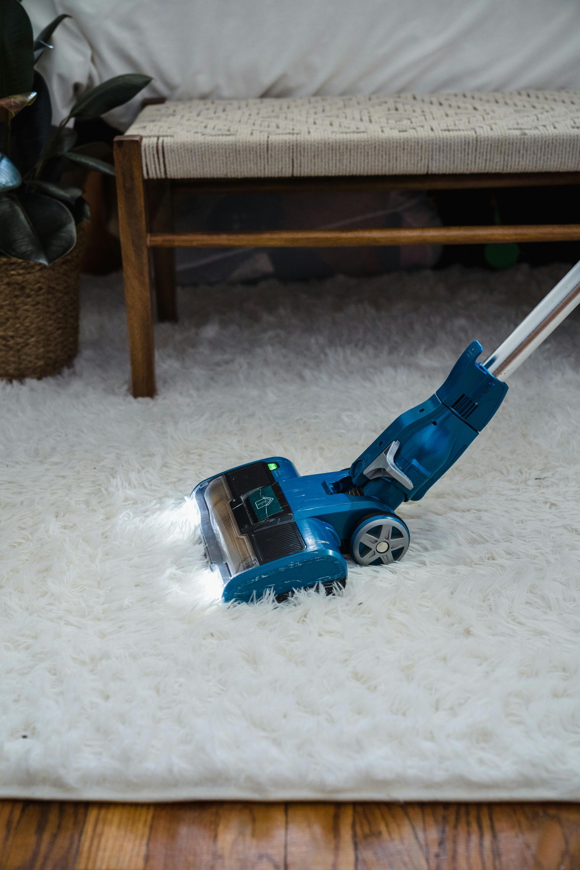 A teal vacuum cleaner with LED headlights cleans a plush white rug in a room with a wooden bench and flooring.