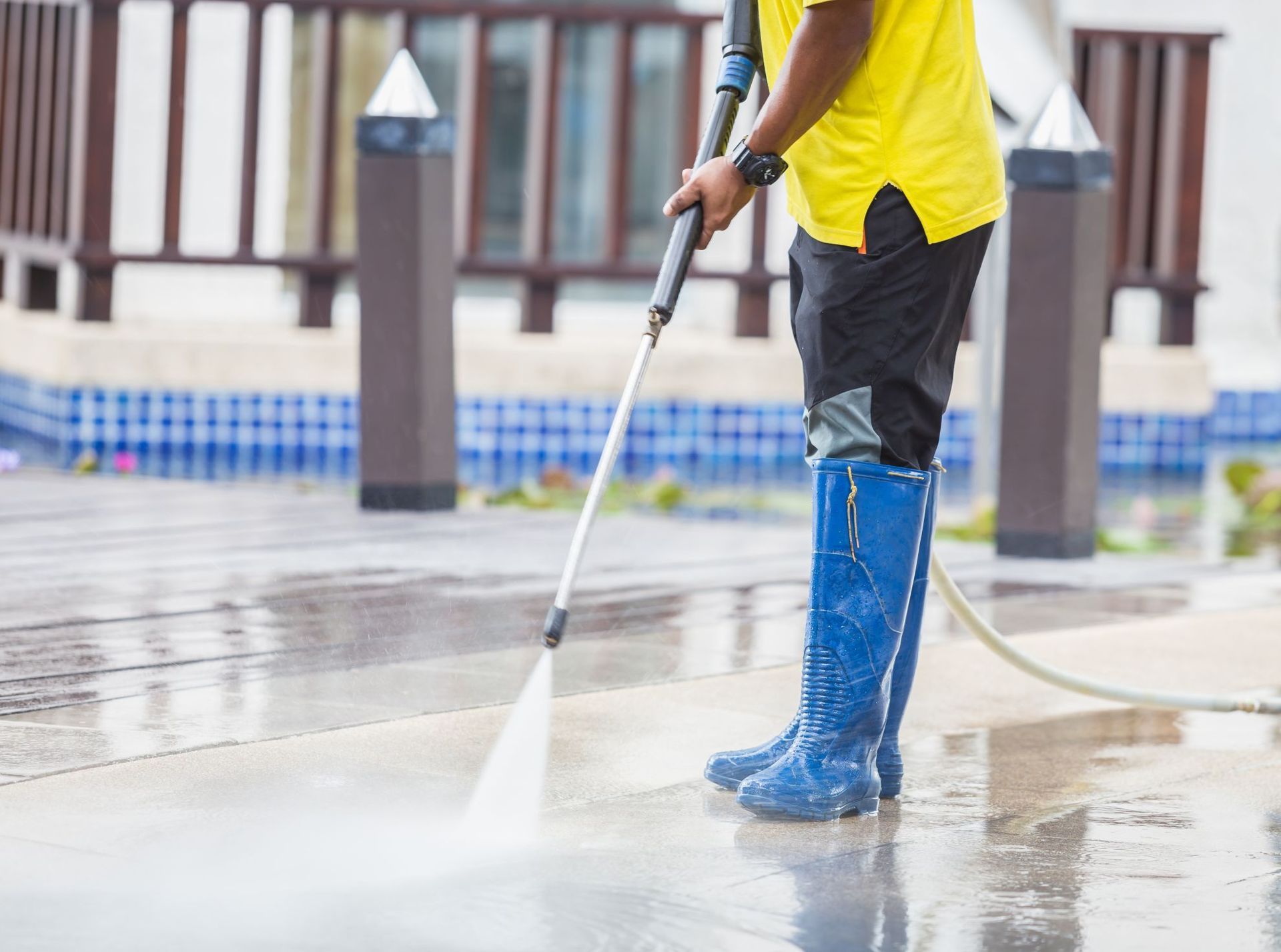 A person in a yellow shirt and blue boots power washes a wet, wooden deck near a swimming pool.