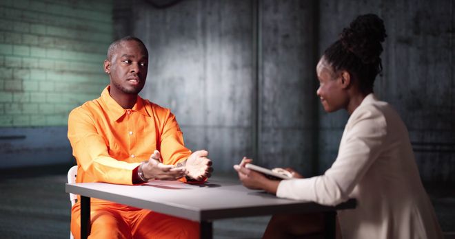A man in an orange jumpsuit is sitting at a table talking to a woman.