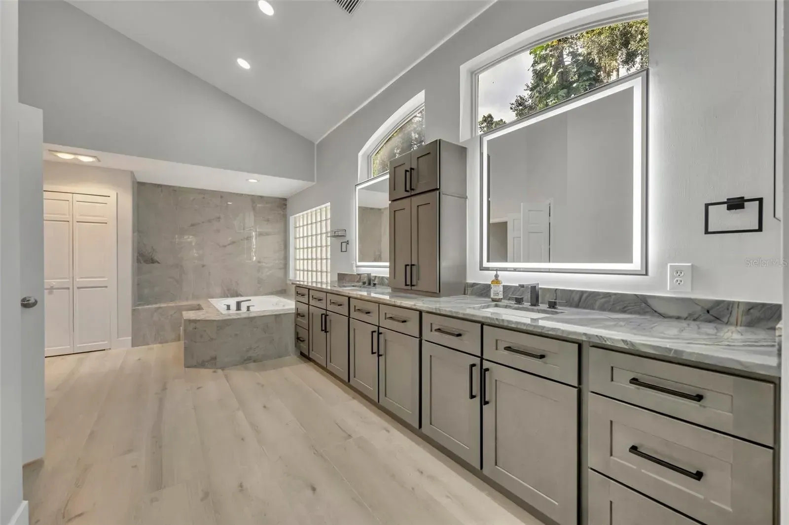 Modern bathroom with freestanding tub, gray vanity, large mirrors, and skylight windows.